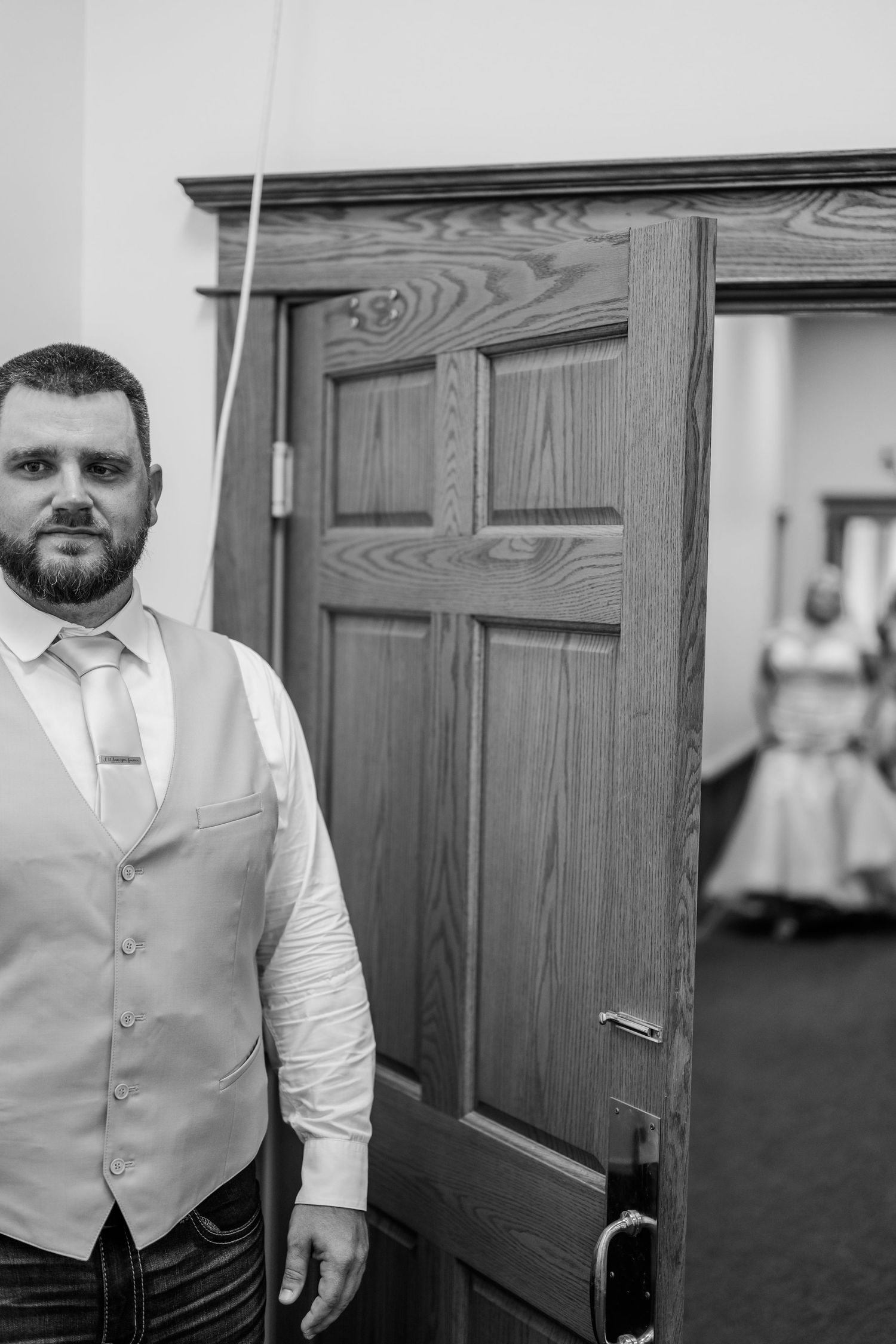 A groomsman in a light vest and white dress shirt stands near wooden doors in a black and white photograph.