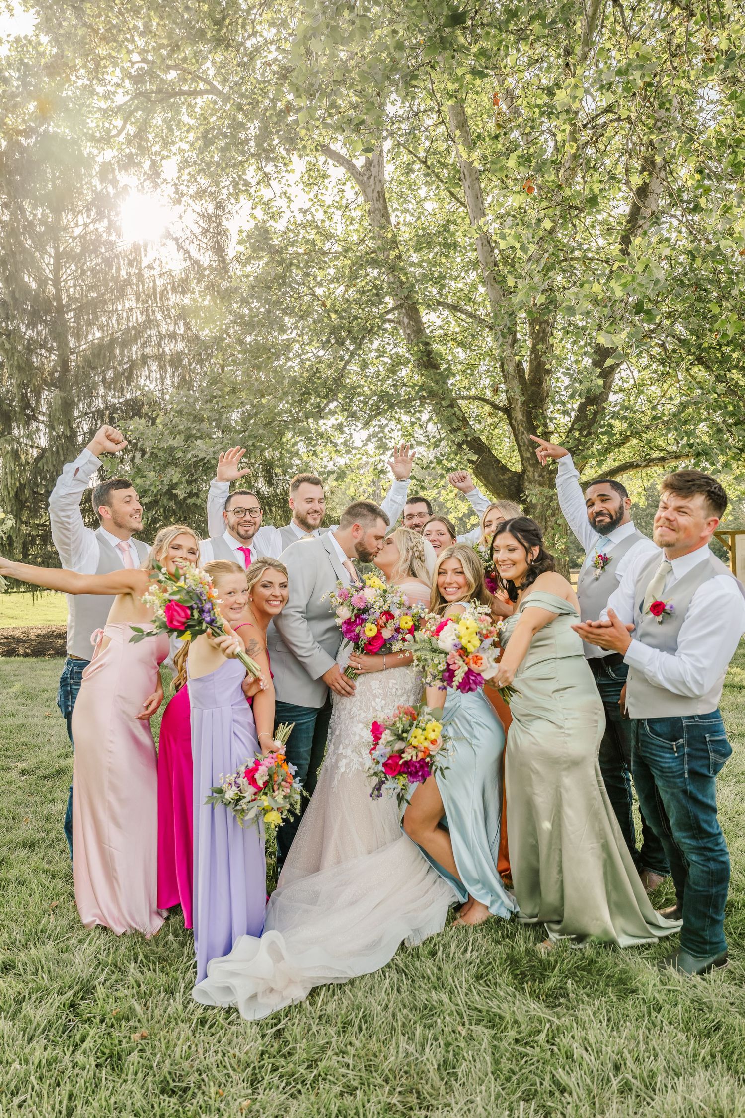 Wedding party in pastel attire poses playfully on grassy lawn during outdoor summer celebration.