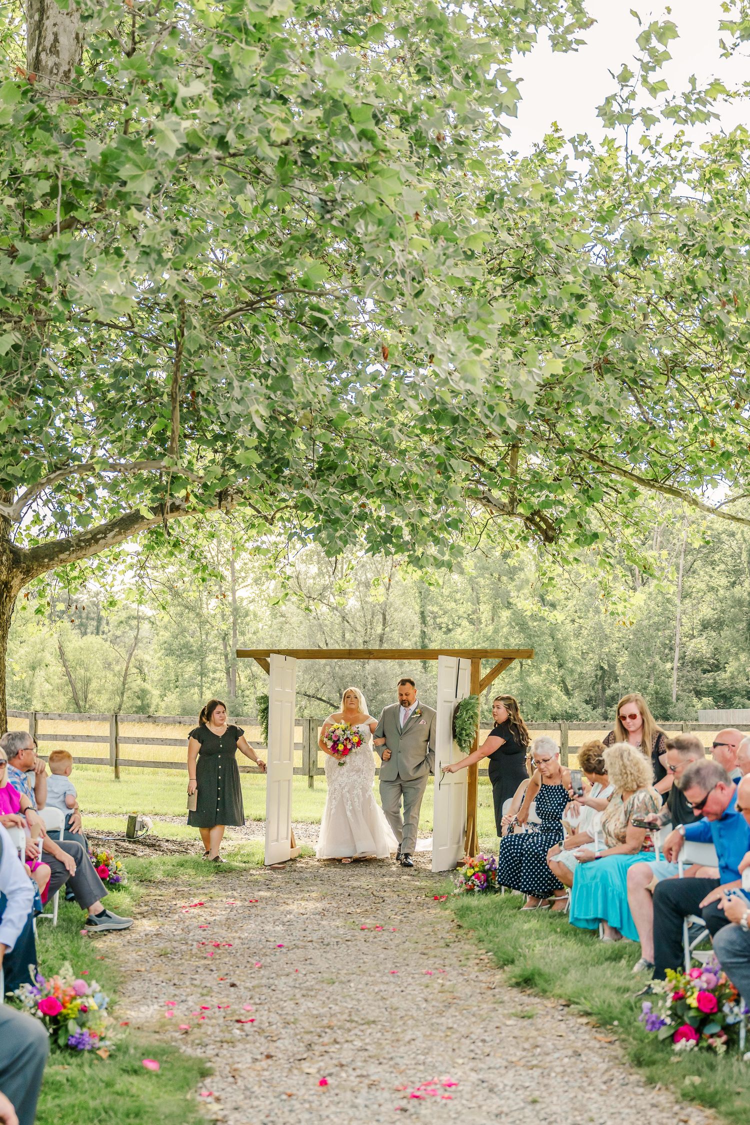 Outdoor wedding ceremony setup with guests seated under trees and scattered rose petals on a natural path.