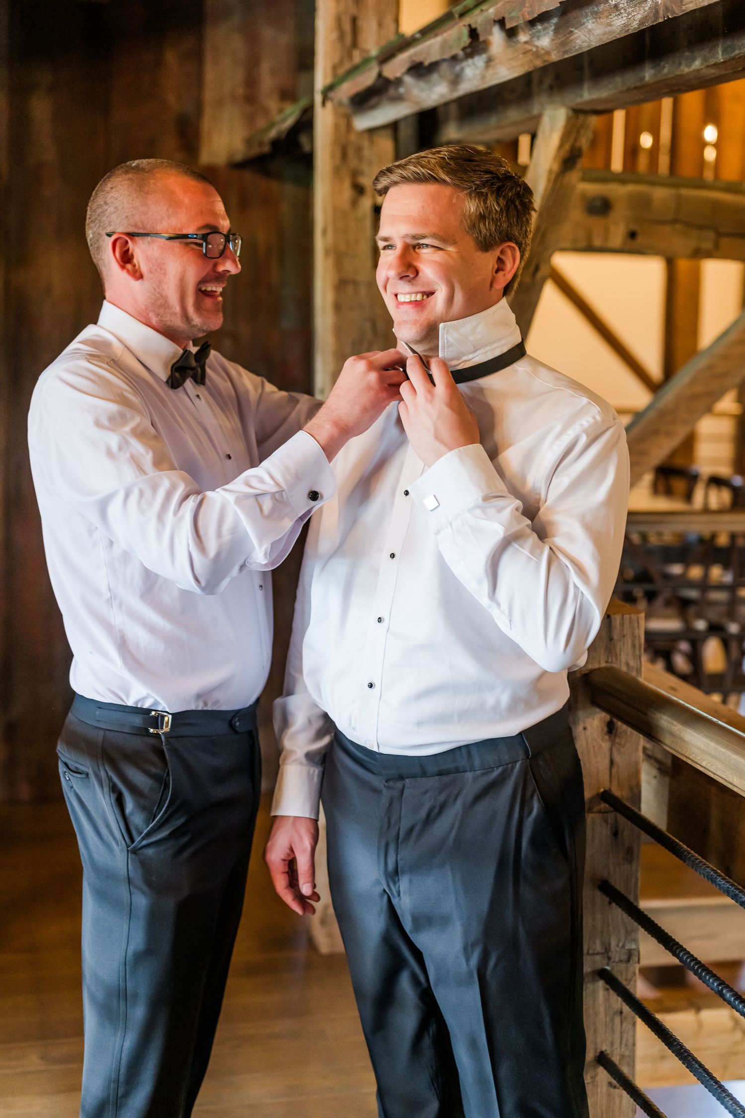 Two people in formal white dress shirts and bow ties share a special moment while adjusting formal attire in a rustic setting.