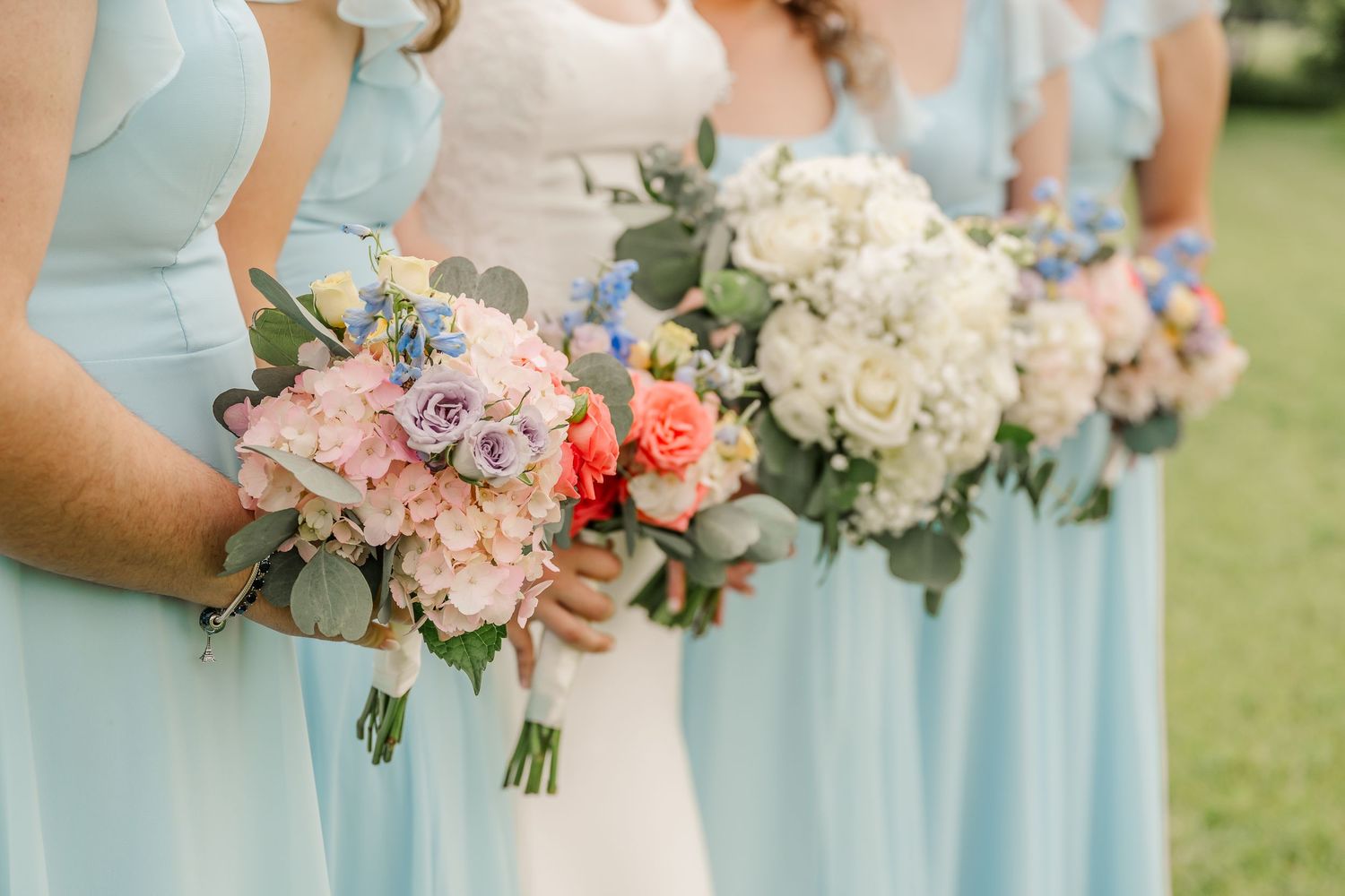 Row of bridesmaids in light blue dresses holding pink and white floral bouquets at an outdoor wedding.