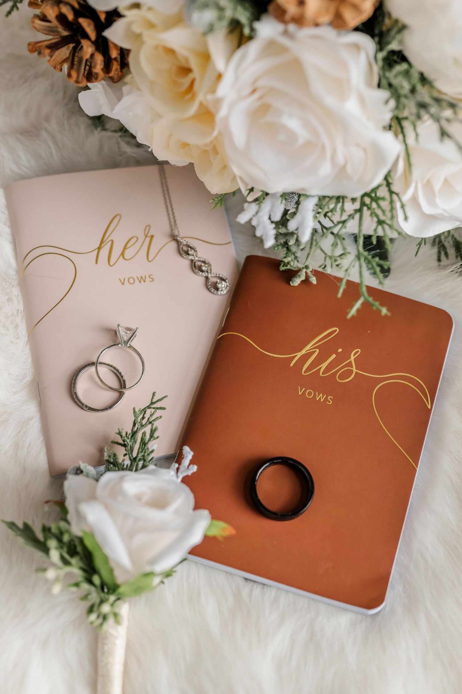 Wedding vow books in brown and beige with rings and white floral decorations arranged on white fabric.