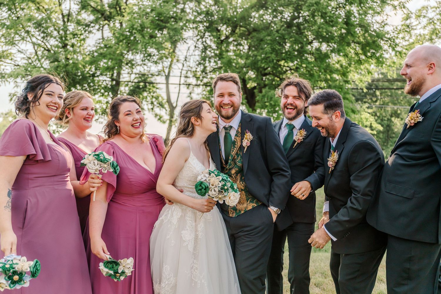 Wedding party in pink dresses and dark suits laugh together in a candid outdoor moment.