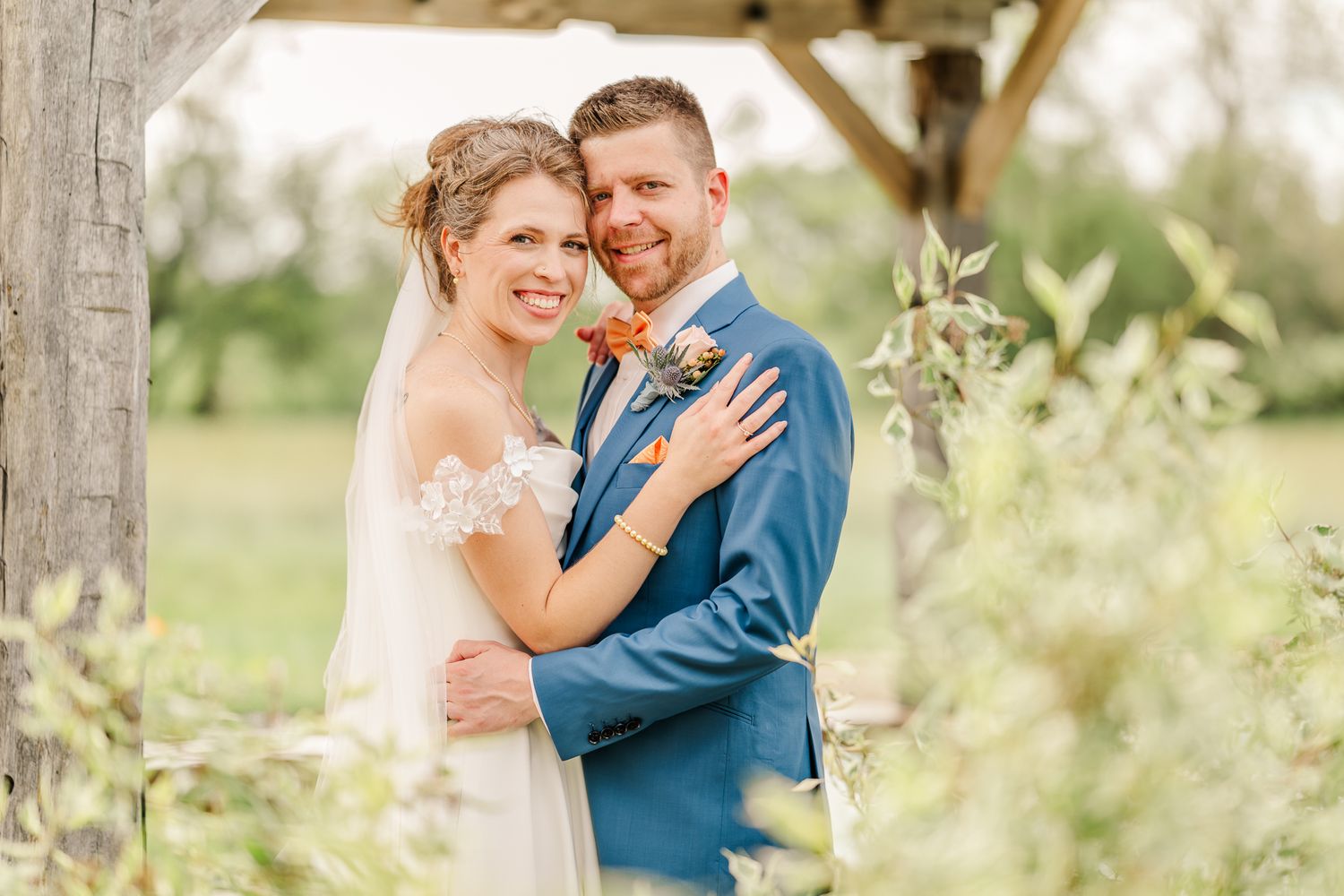 Bride & groom embrace under wooden arbor, she in lace gown with veil, he in blue suit with floral boutonniere.The Old Barns at Dry Run Farms