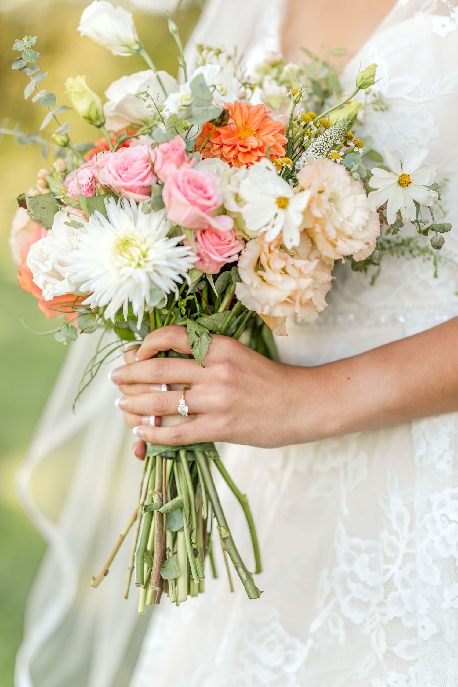 Bride in a lace wedding gown holds a pastel bridal bouquet of pink roses, white dahlias, orange blooms and greenery.