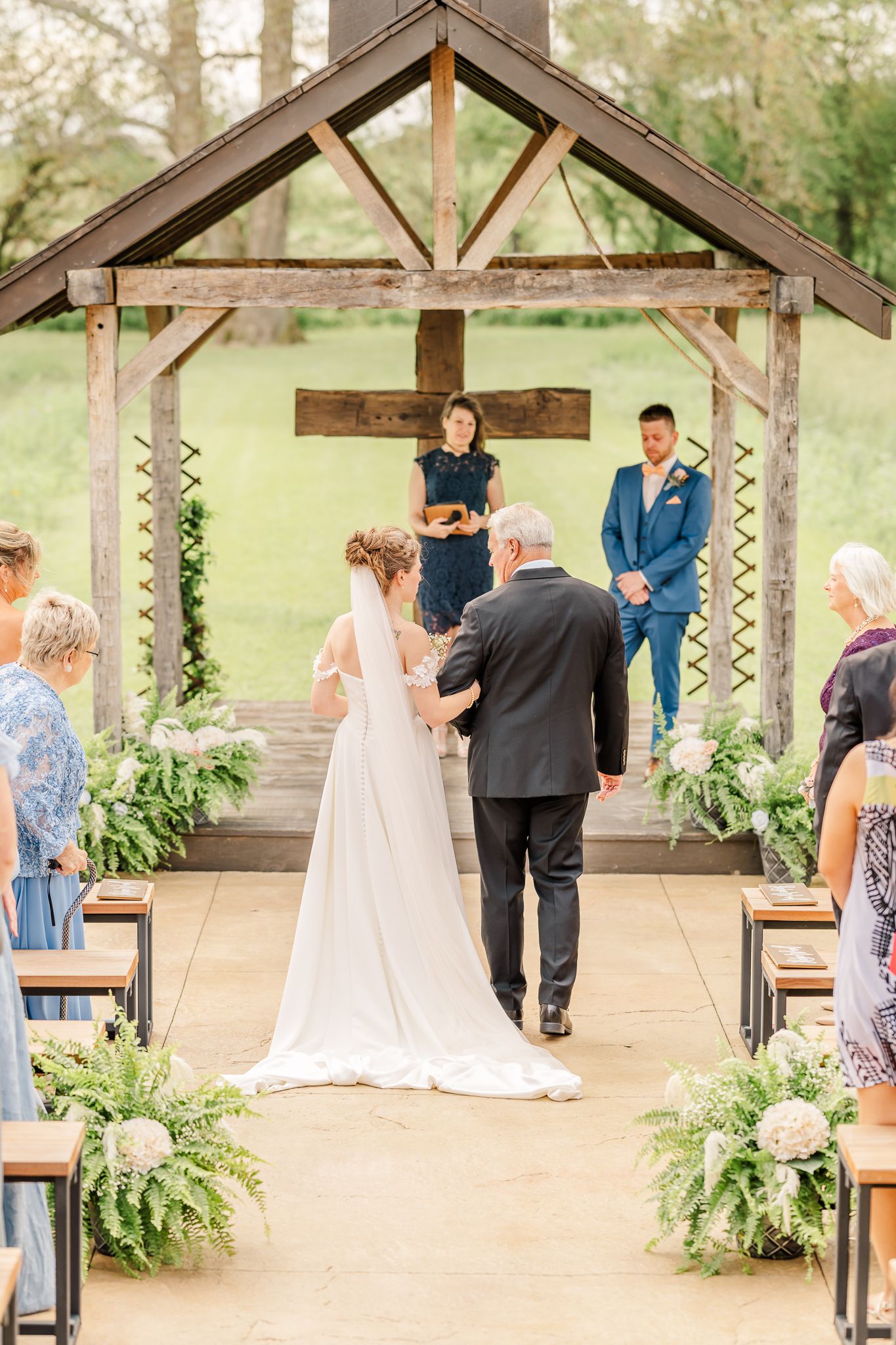 Bride walks down the aisle with her father toward the groom at an outdoor chapel wedding ceremony with wooden cross.