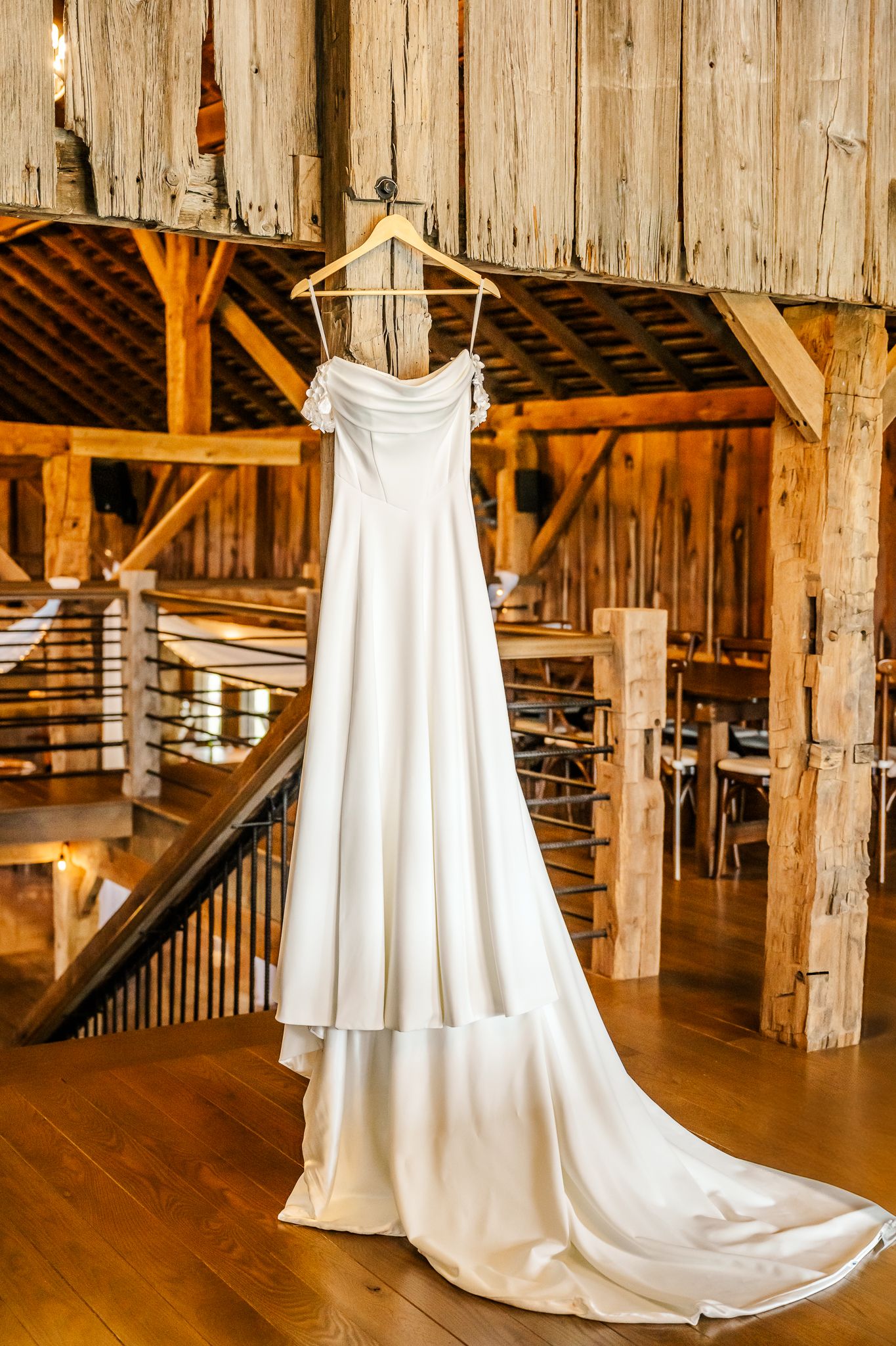 An elegant white wedding gown with a long train hangs on a wooden hanger inside a rustic barn venue.The Old Barns at Dry Run Farms.