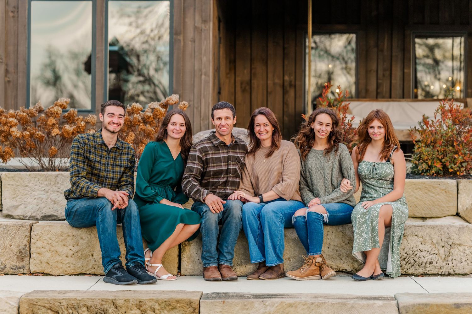Six young adults smiling and sitting together on a stone bench outside a rustic building in autumn. The Old Barns at Dry Run Farms.