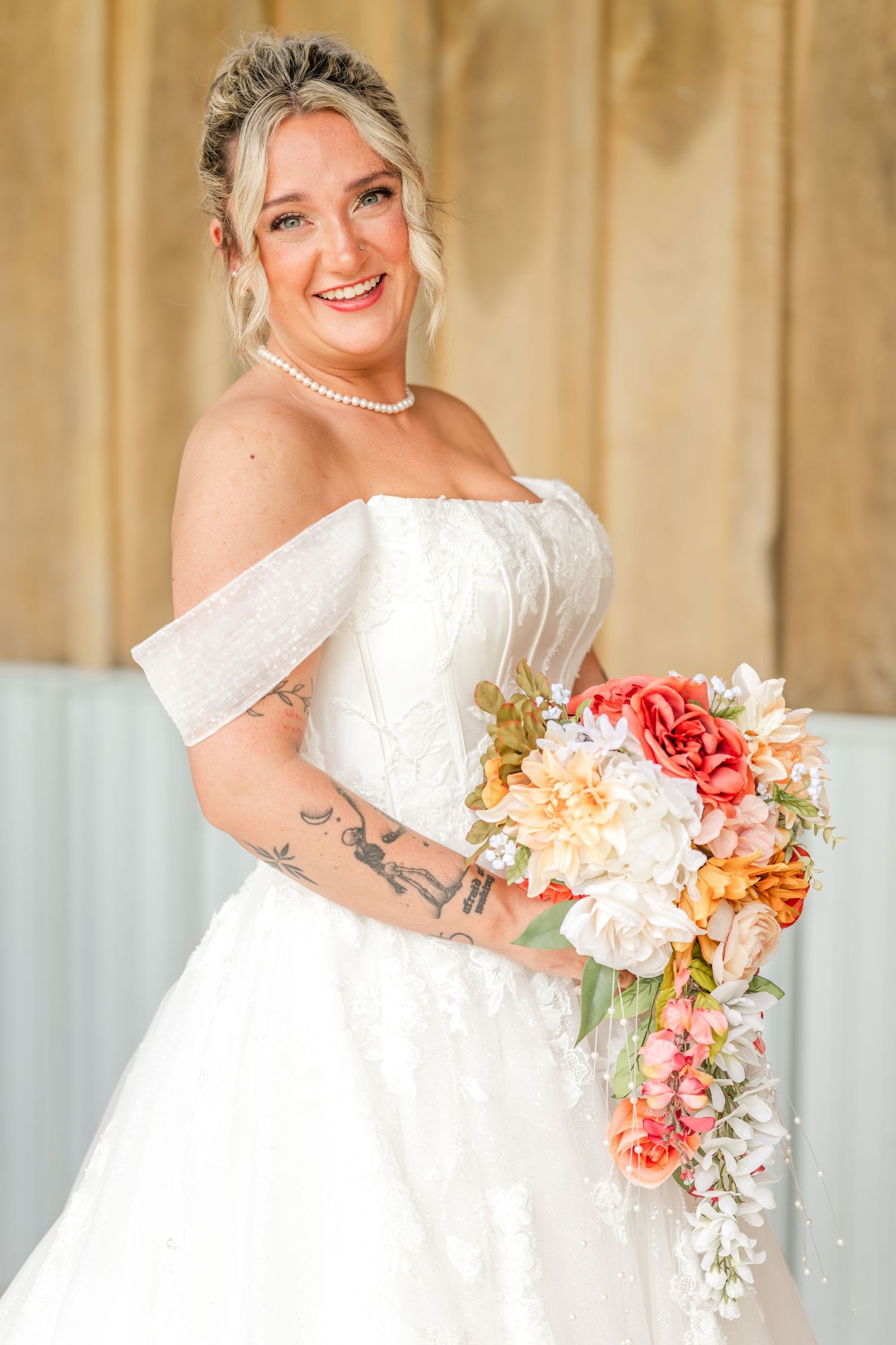 Smiling bride in an off-the-shoulder white lace wedding gown holding a coral, peach and white cascading floral bouquet.