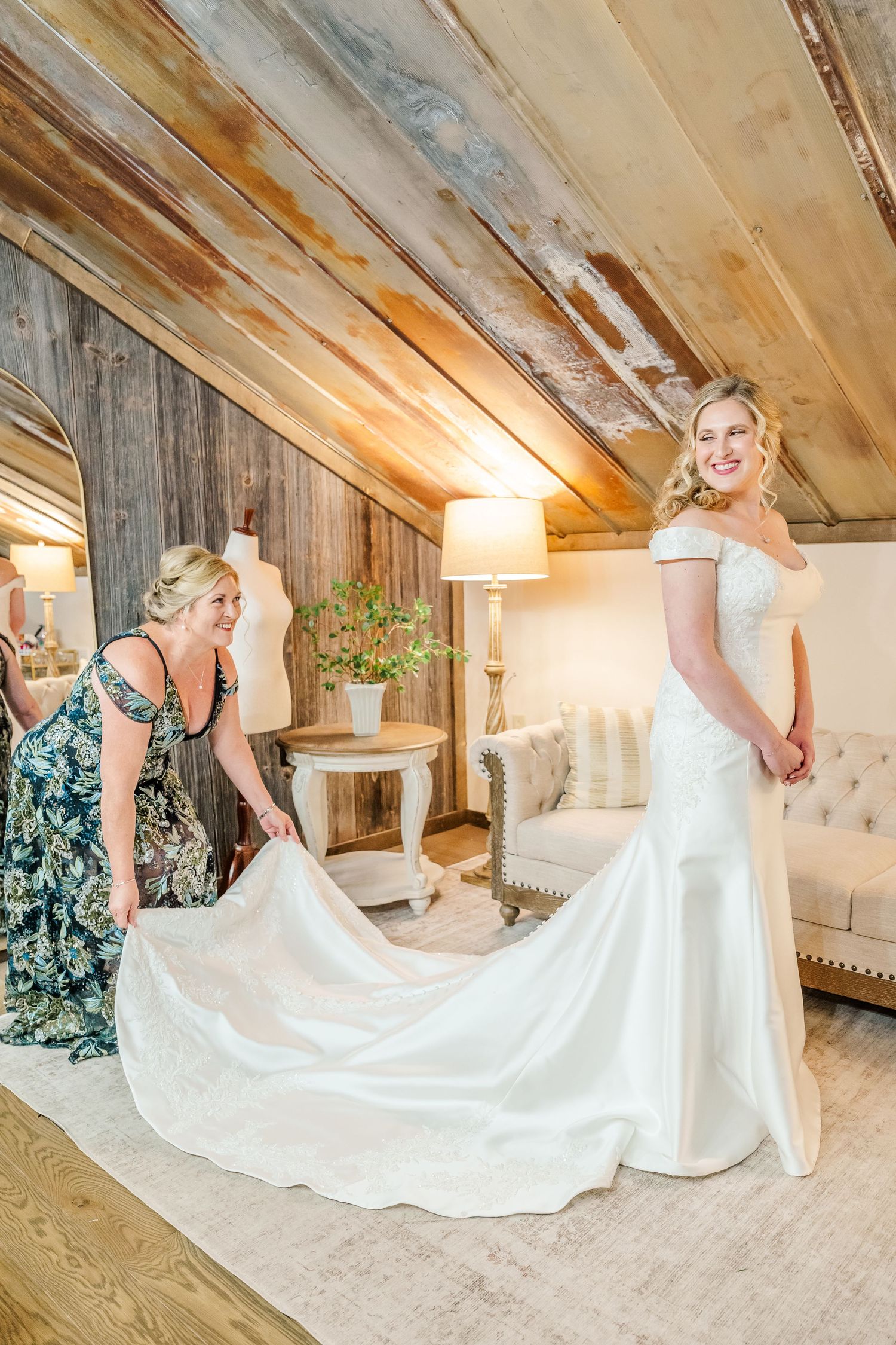 A person in an elegant off-shoulder white wedding gown has their train adjusted in a rustic wooden room.The Old Barns at Dry Run Farms