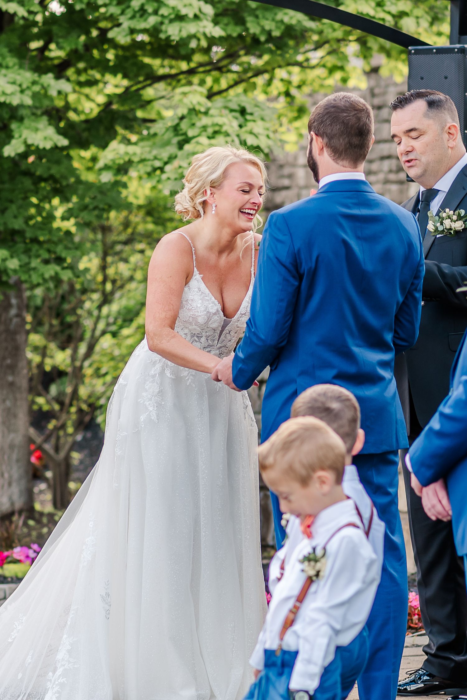 A joyful wedding celebration outdoors with guests in formal attire gathered under green summer foliage.