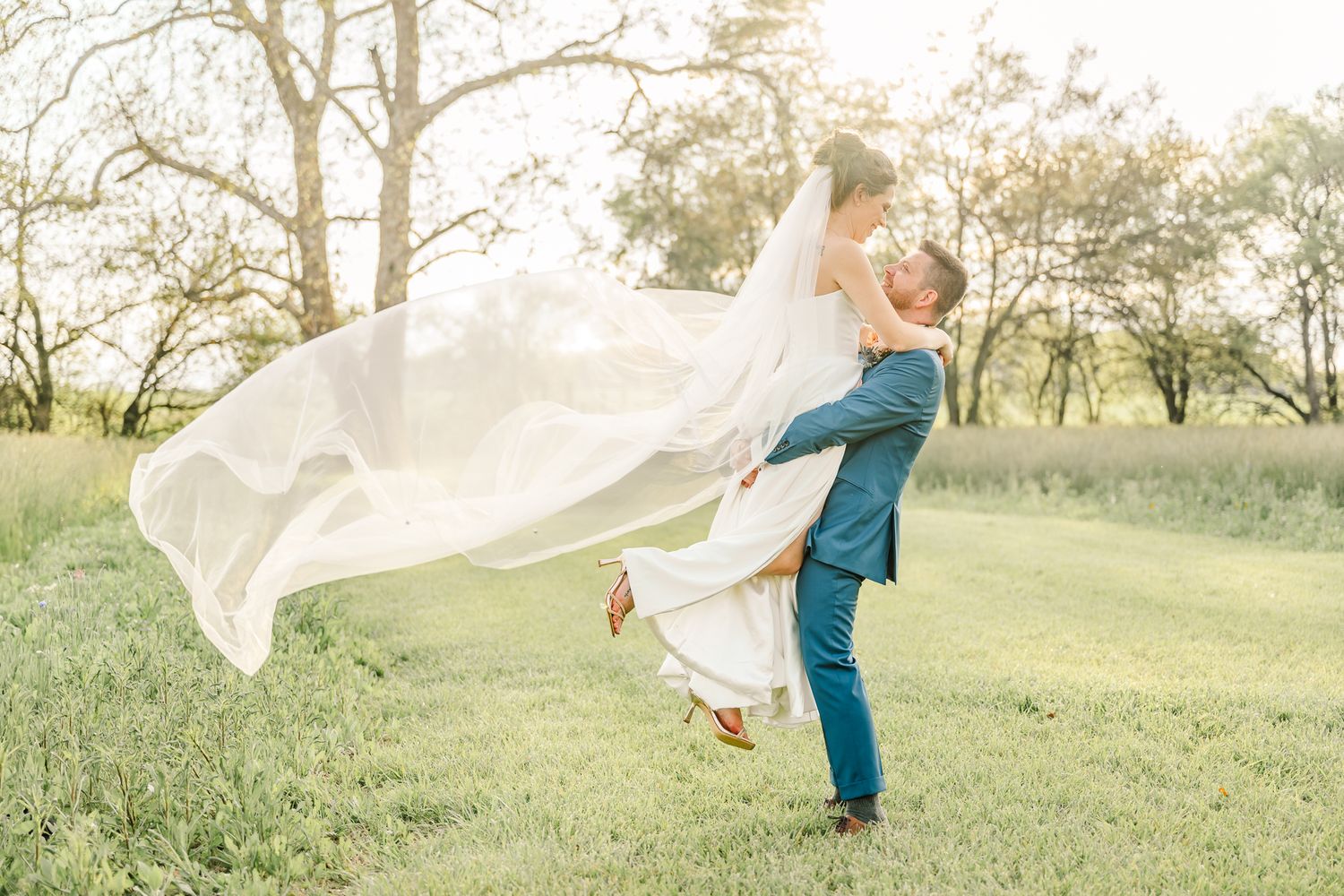 Groom lifting bride in white gown and flowing veil in a sunlit green meadow during wedding photos.
