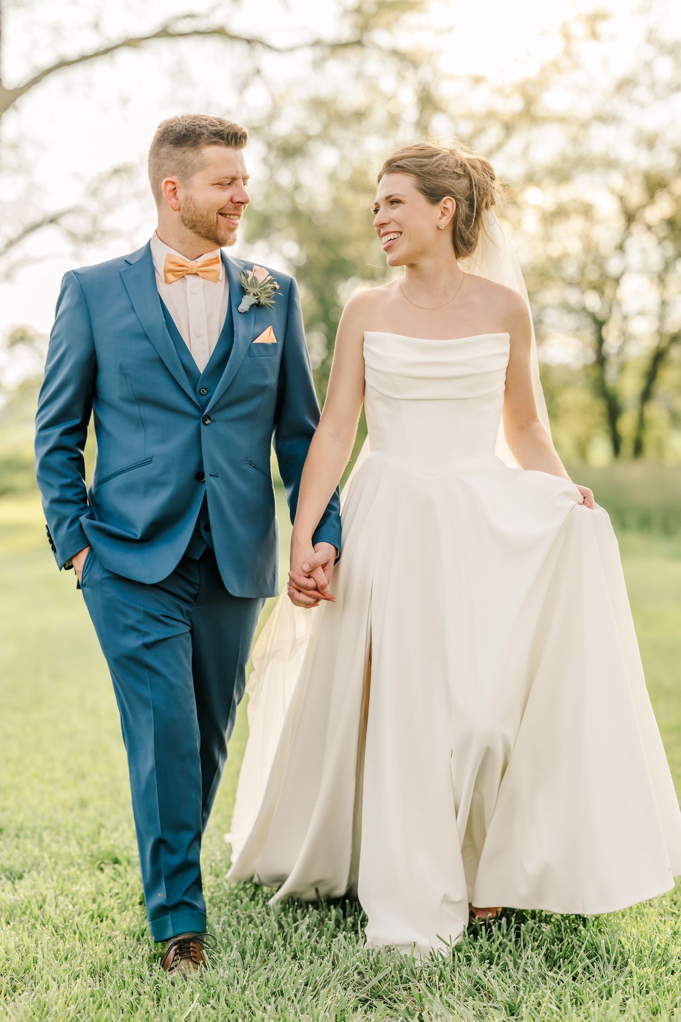 Smiling bride and groom walk hand in hand outdoors, she in strapless gown, he in blue suit with peach bow tie.The Old Barns at Dry Run Farms
