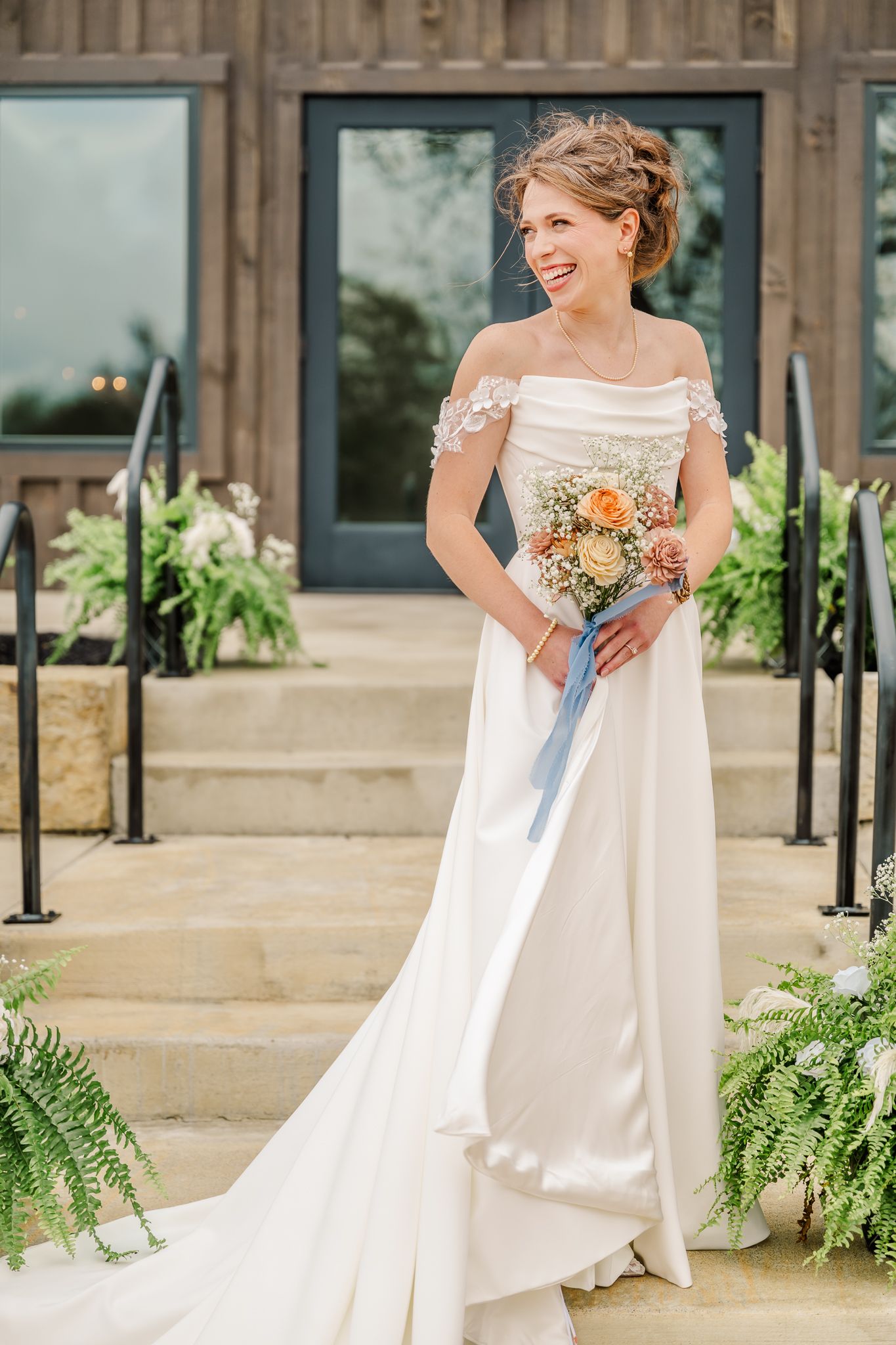 Smiling bride in an off-shoulder white satin gown holding a peach and blush bouquet on stone steps.The Old Barns at Dry Run Farms in Ohio.