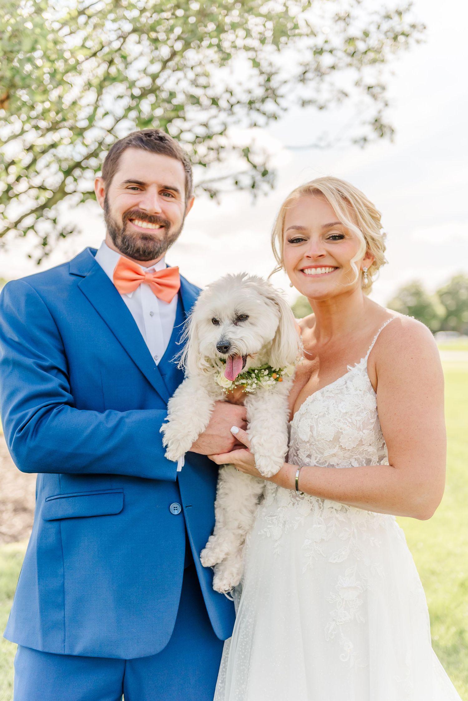 A couple in wedding attire holds their small white dog wearing a floral collar during outdoor wedding photos.