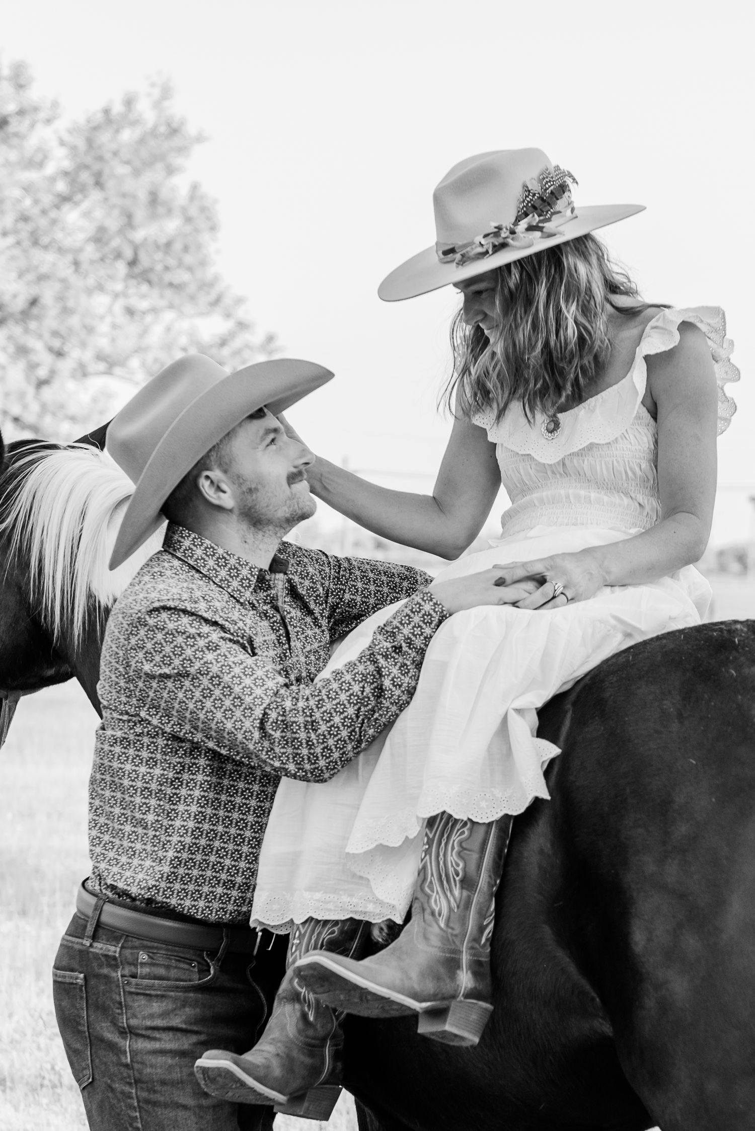 A romantic black and white western engagement photo with a couple in cowboy attire posing with their horse outdoors.