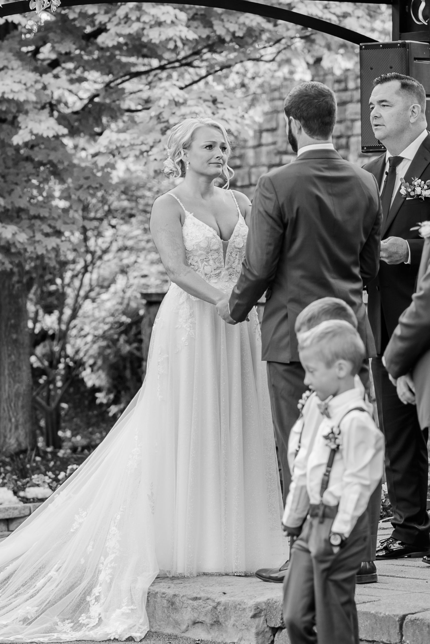 A black and white wedding ceremony scene outdoors with guests gathered under trees.