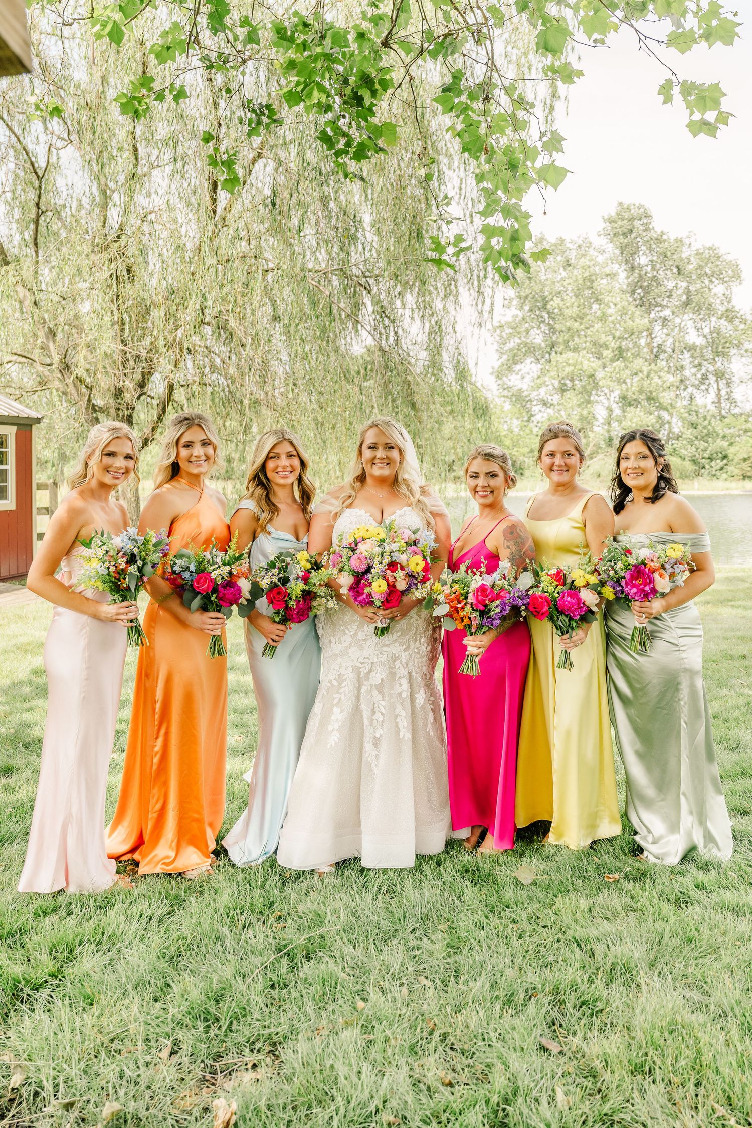 Bridesmaids in colorful satin dresses hold vibrant floral bouquets while posing under a willow tree at an outdoor wedding.