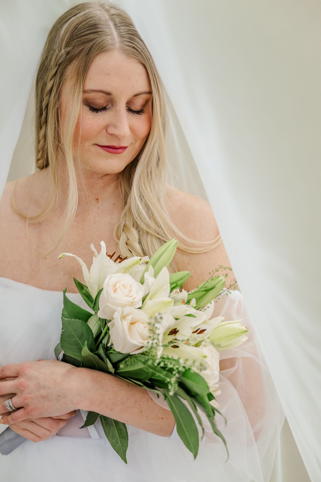 Blonde bride in white gown holds elegant bouquet of white roses and lilies, wearing veil and braided hair.