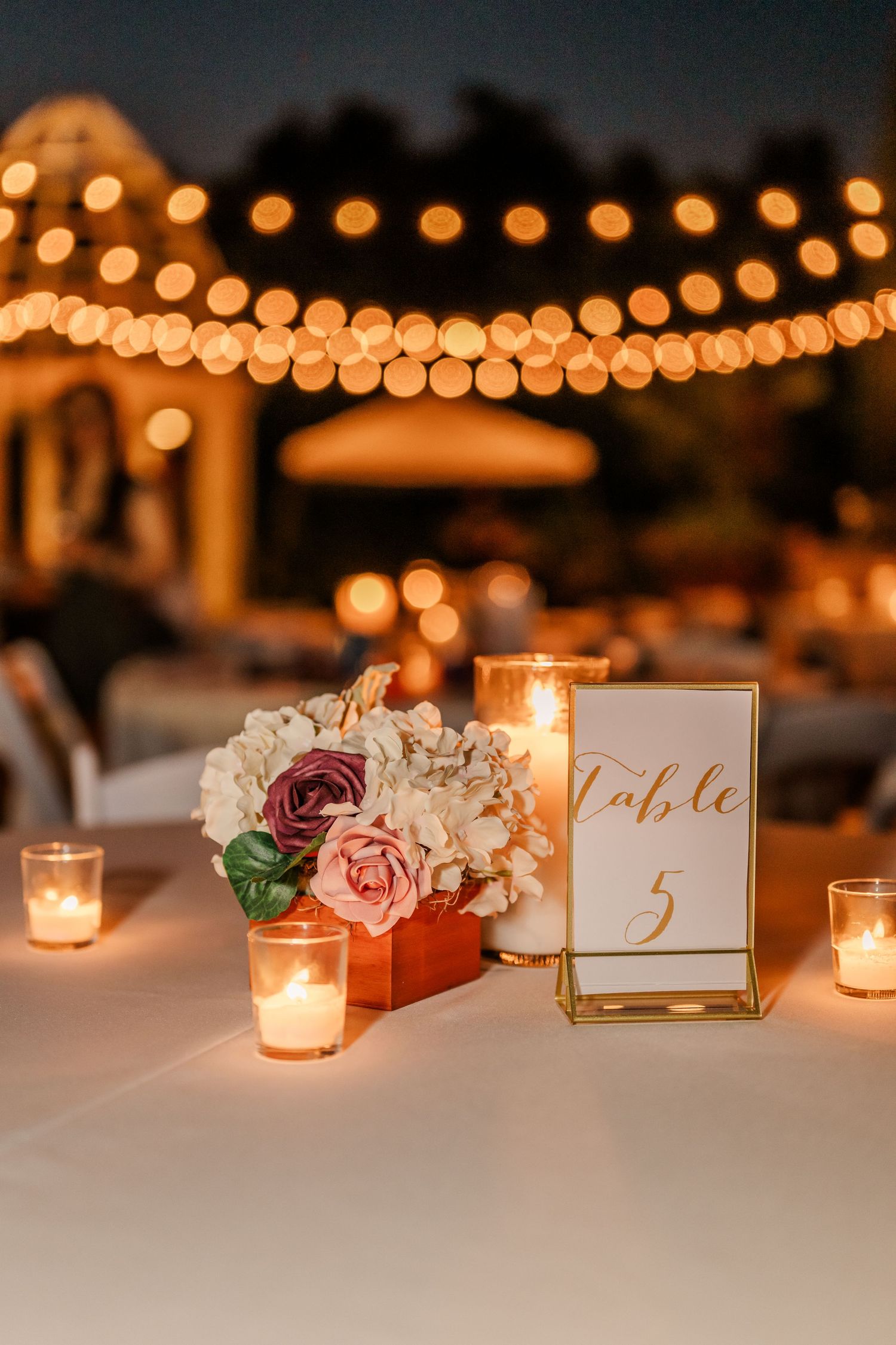Romantic table settings with candlelight, floral arrangements, and gold table numbers under string lights.