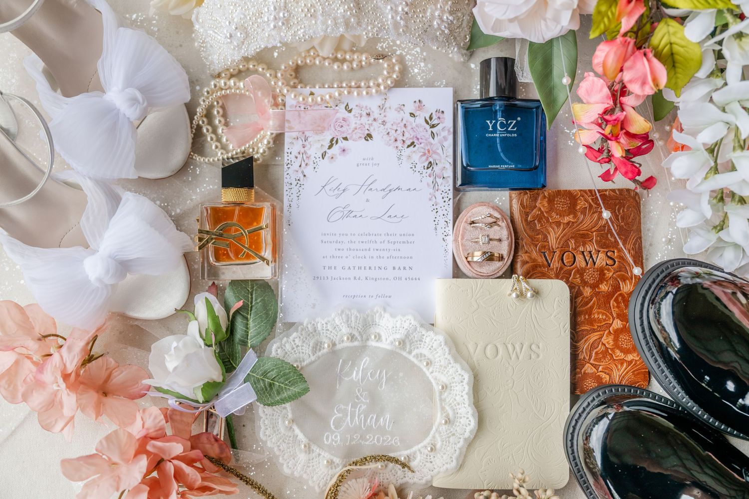 Flat lay of wedding day details with an invitation suite, vow books, perfume bottles, pink flowers, pearls, a veil and black dress shoes.