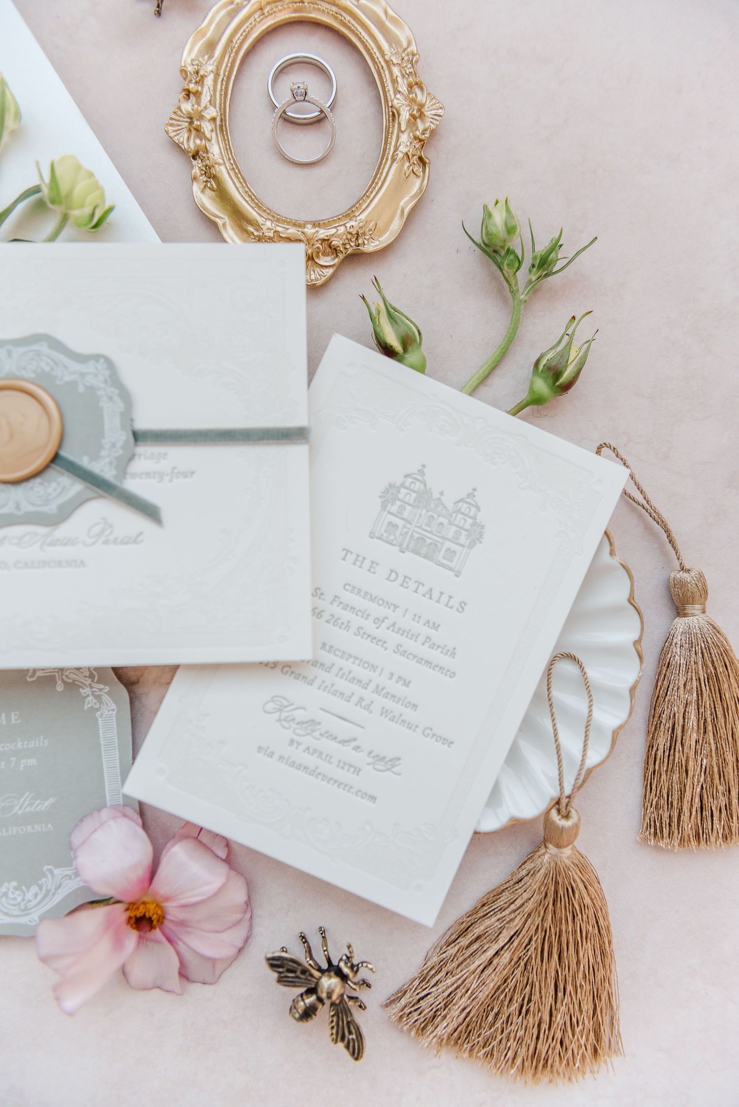 Elegant wedding invitation suite with beige tassels, ornate gold frames, and delicate pink flowers on a neutral background.