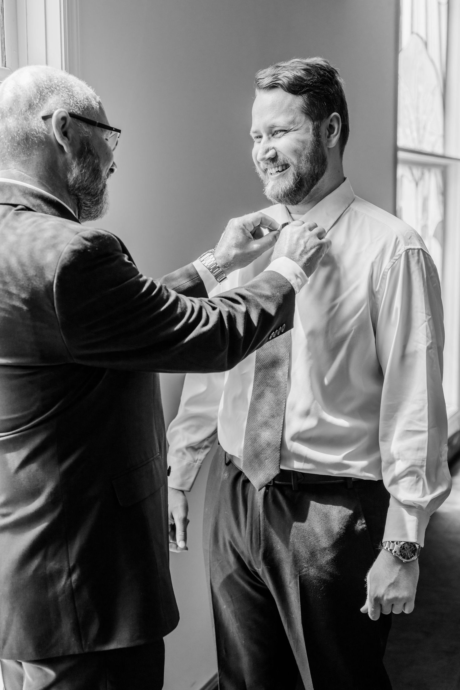 Black and white photo showing a man helping another adjust his tie before a formal event.
