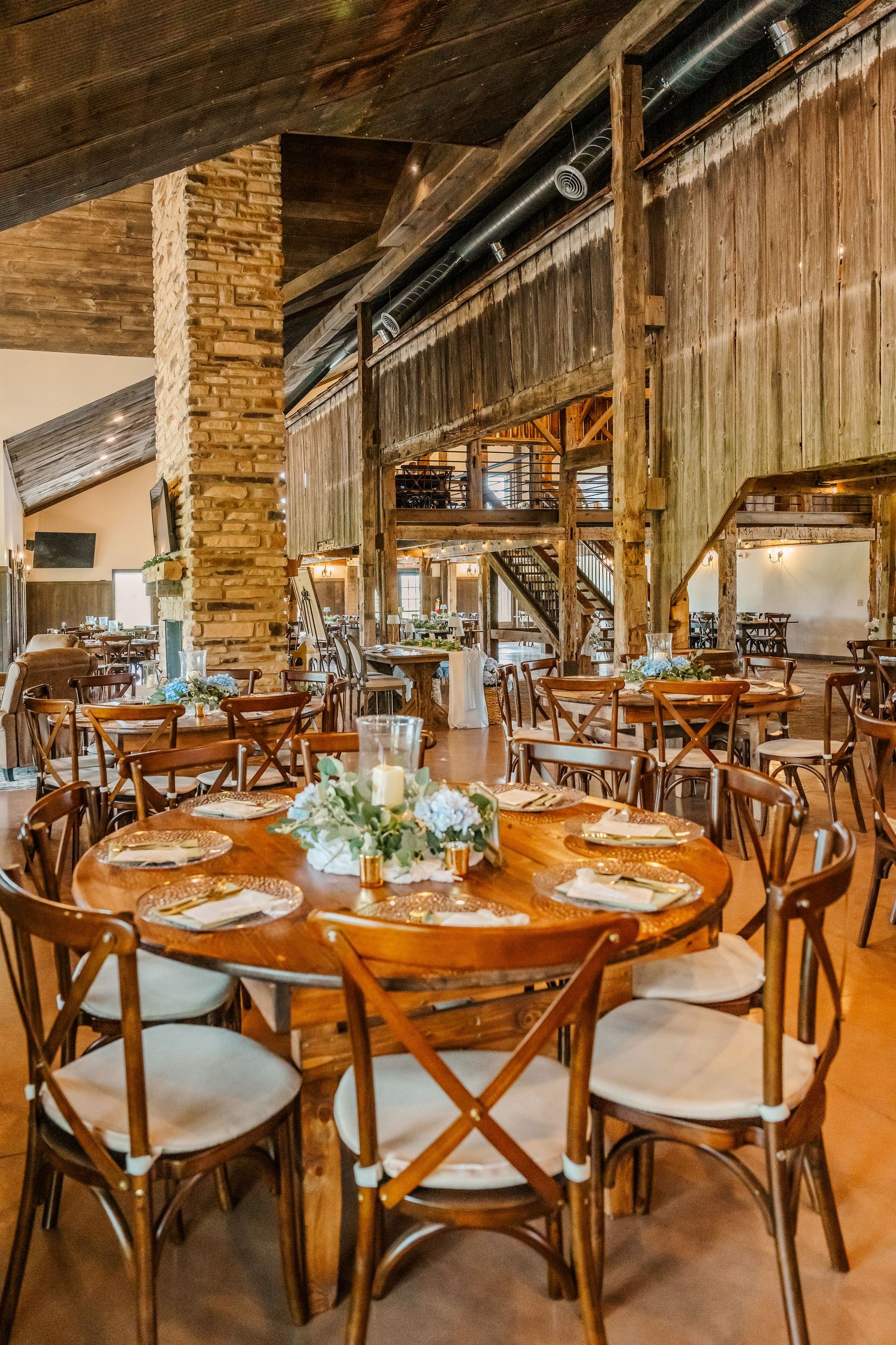 Rustic wedding reception venue with wooden cross-back chairs and round tables decorated with elegant centerpieces.
