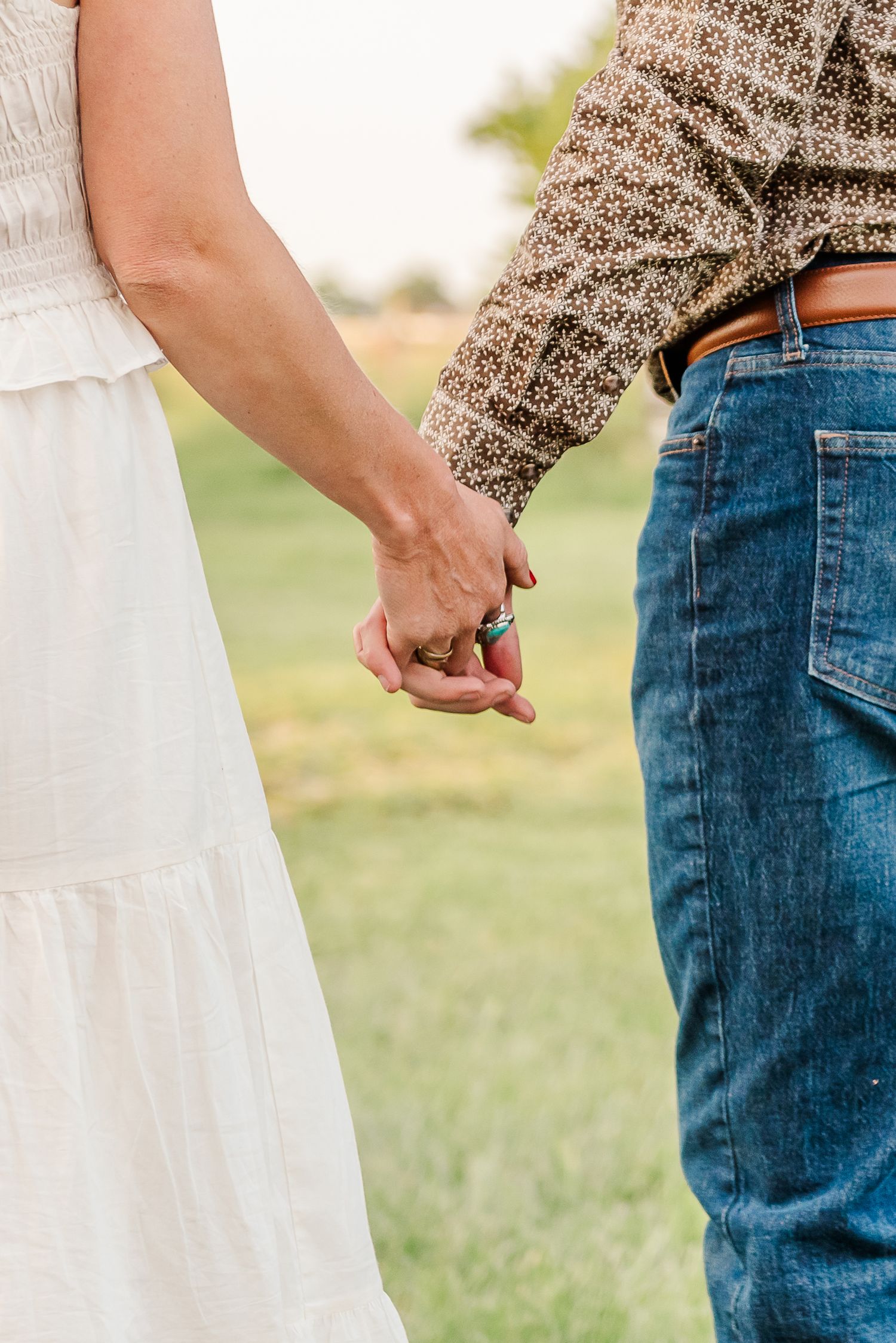 A couple holds hands in a grassy field, wearing a white dress and brown sweater with blue jeans.