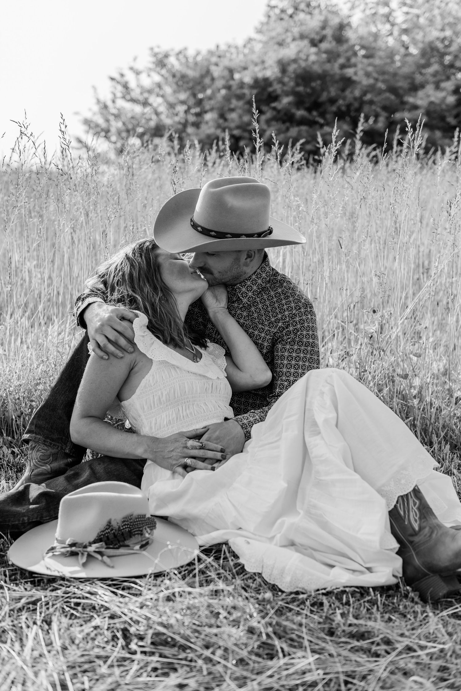 A romantic black and white photograph of a couple in western attire sitting together in a grassy field.