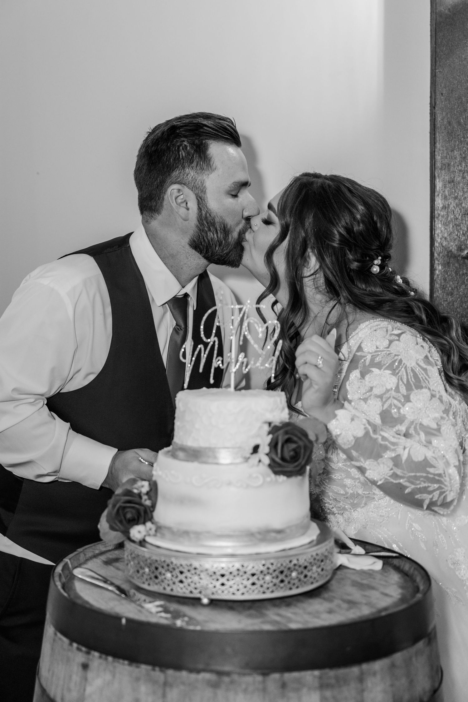 A romantic moment between newlyweds sharing a kiss behind their wedding cake displayed on a rustic barrel.Four Seasons Barn Wedding