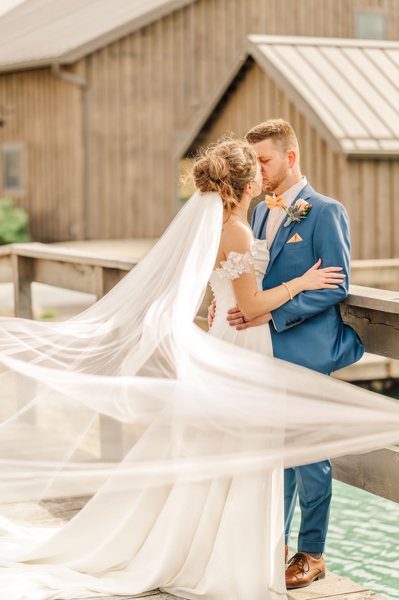 Bride and groom share a romantic kiss on a wooden dock, her veil flowing in the breeze, groom in a blue suit.