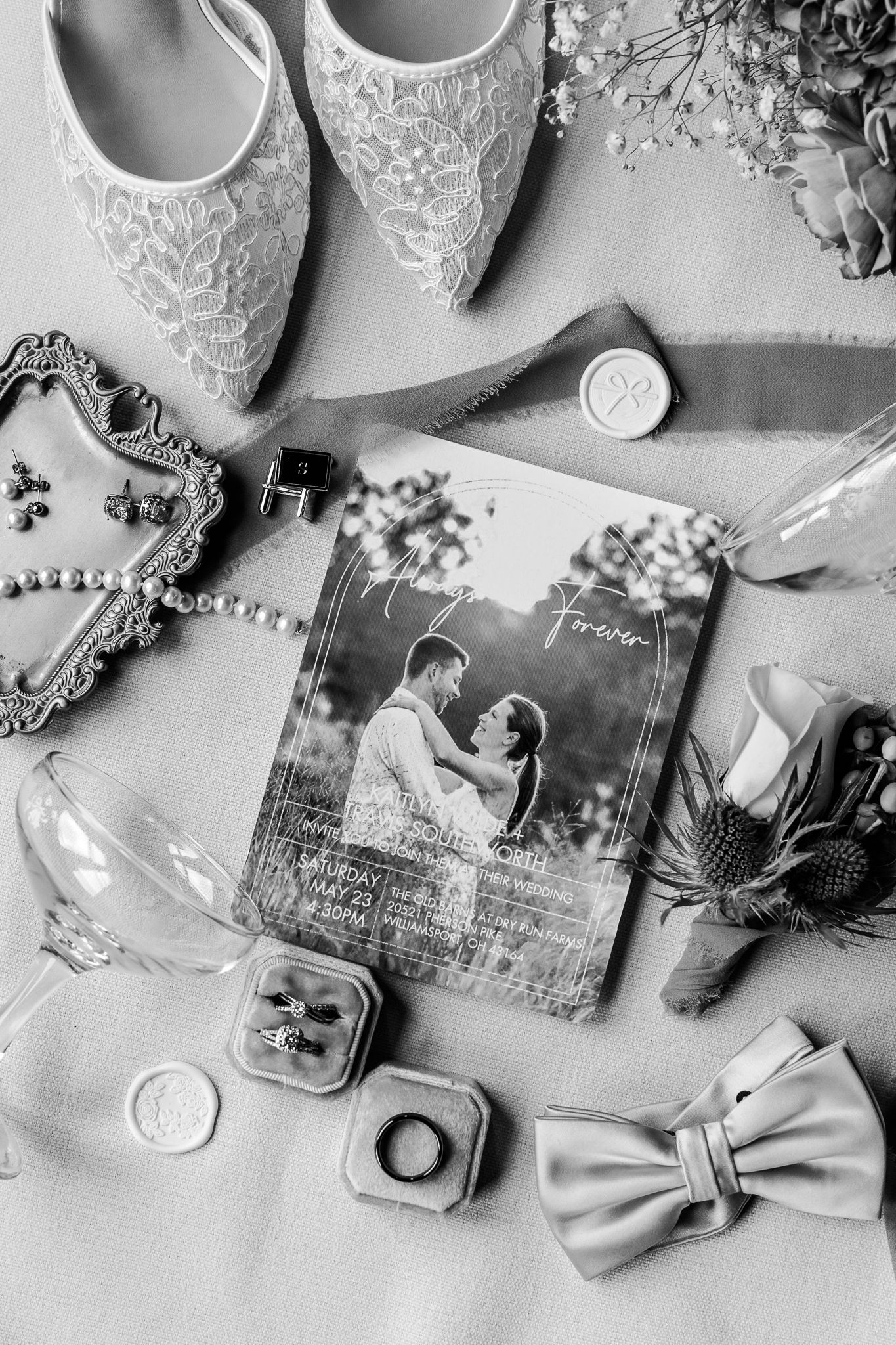 Black and white flat lay of wedding details including lace heels, rings, bow tie, invitation, and floral accents.