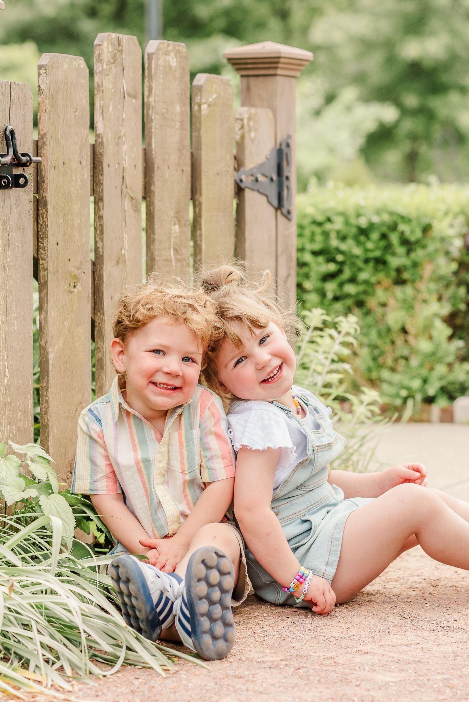 Two young children in casual clothing playfully lounging by a wooden fence in a garden setting.