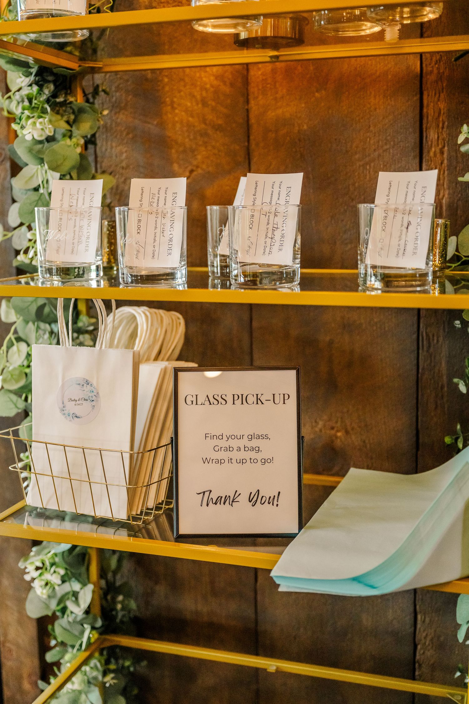 Wedding reception drink station with handwritten signs and glass pickup area decorated with eucalyptus garlands.
