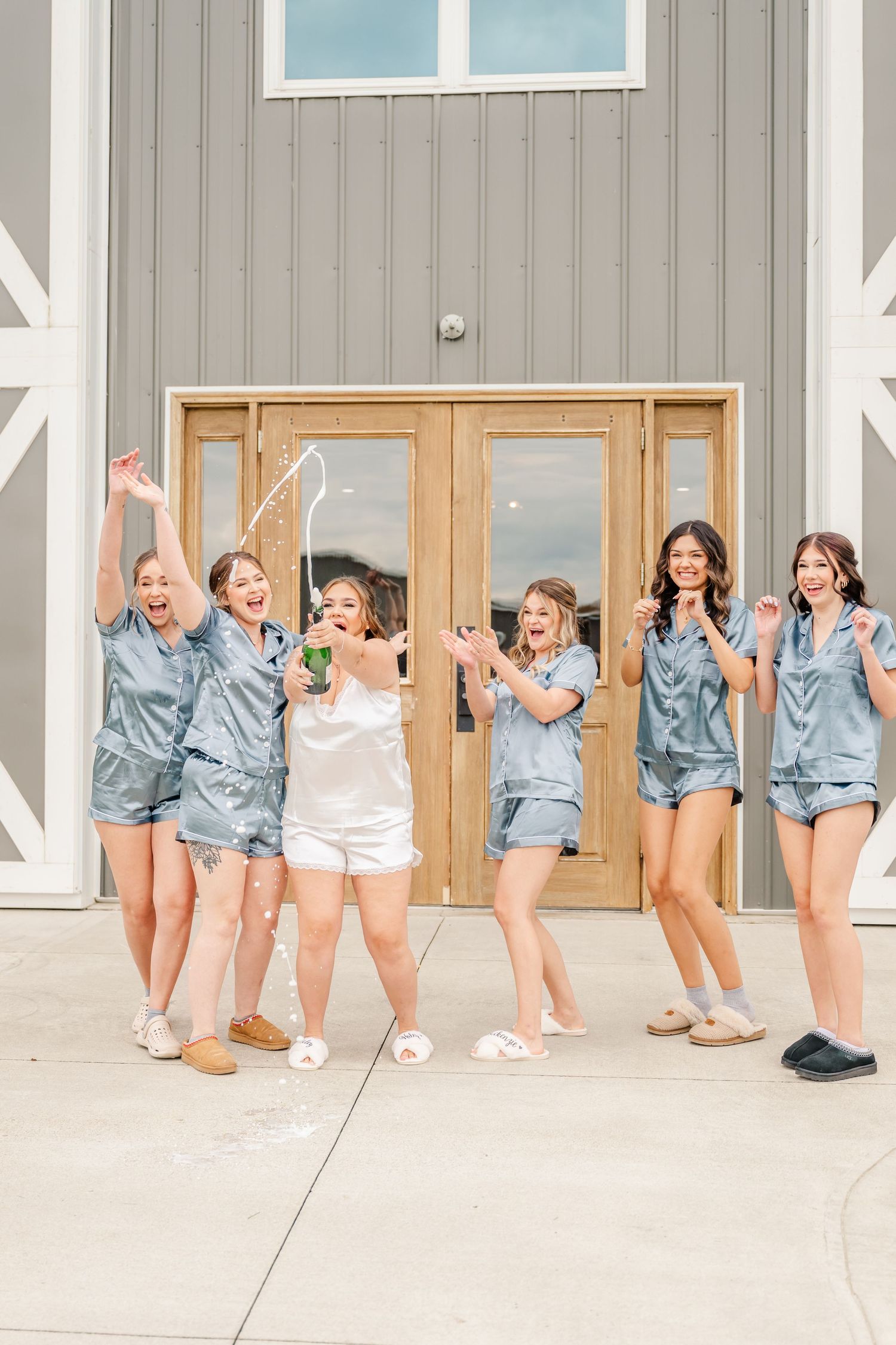 Wedding party in matching blue robes jumping and celebrating outside a modern gray building.Four Seasons Barn Wedding & Event Venue