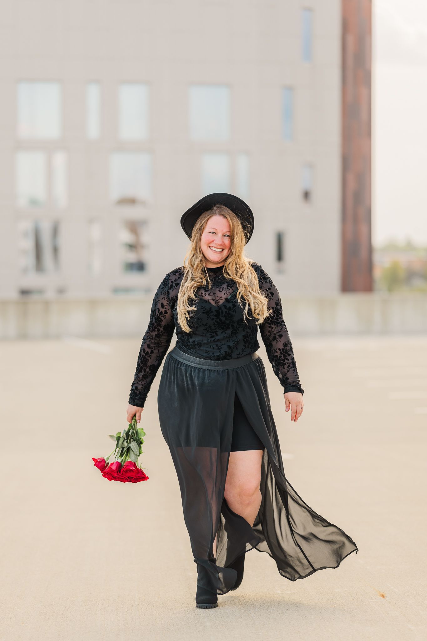 Smiling plus-size woman in black lace outfit and hat holding red roses, walking outdoors near a modern building.