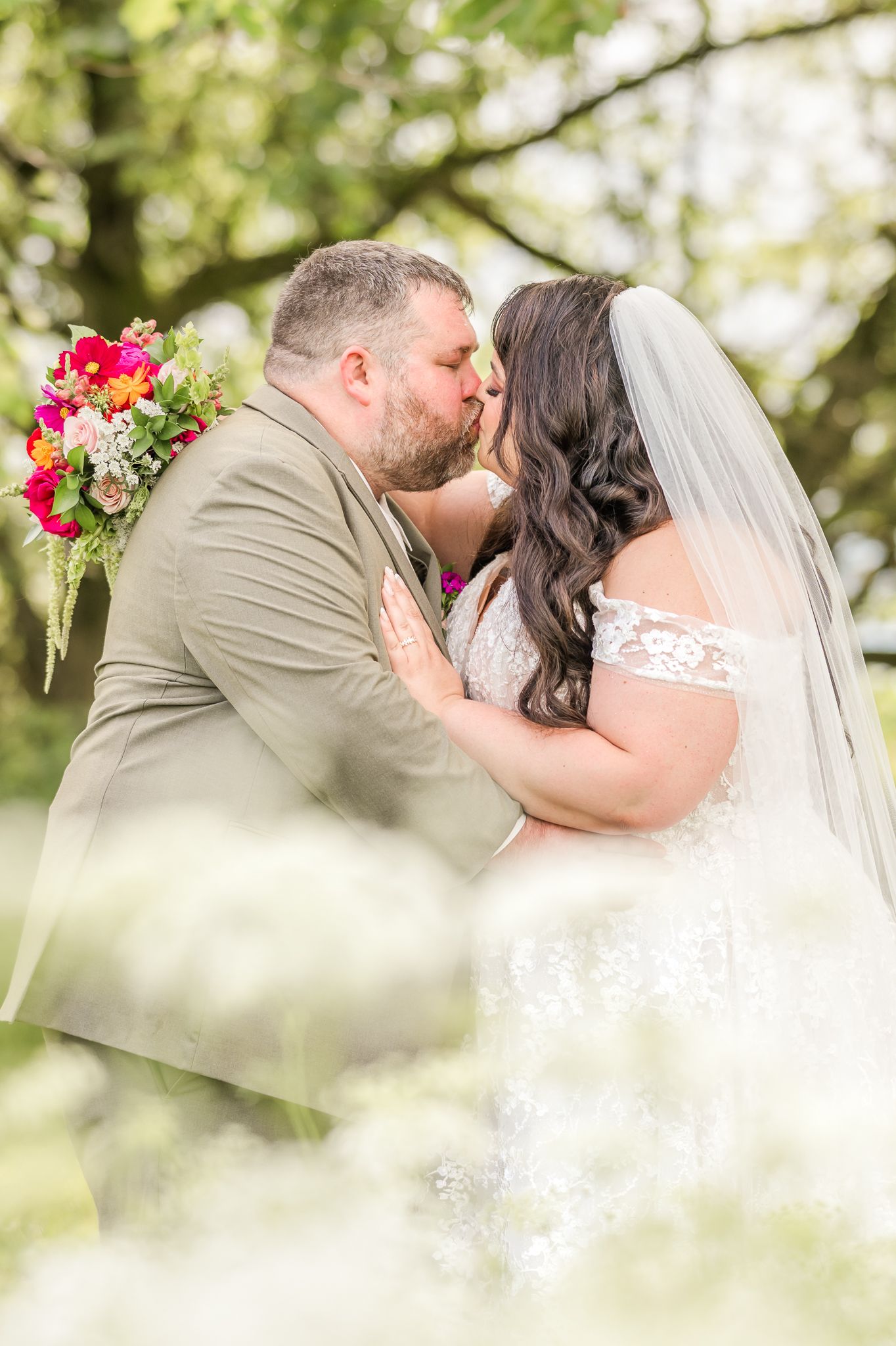Bride and groom share a romantic kiss outdoors, bride in lace gown with veil, groom in tan suit holding colorful bouquet.