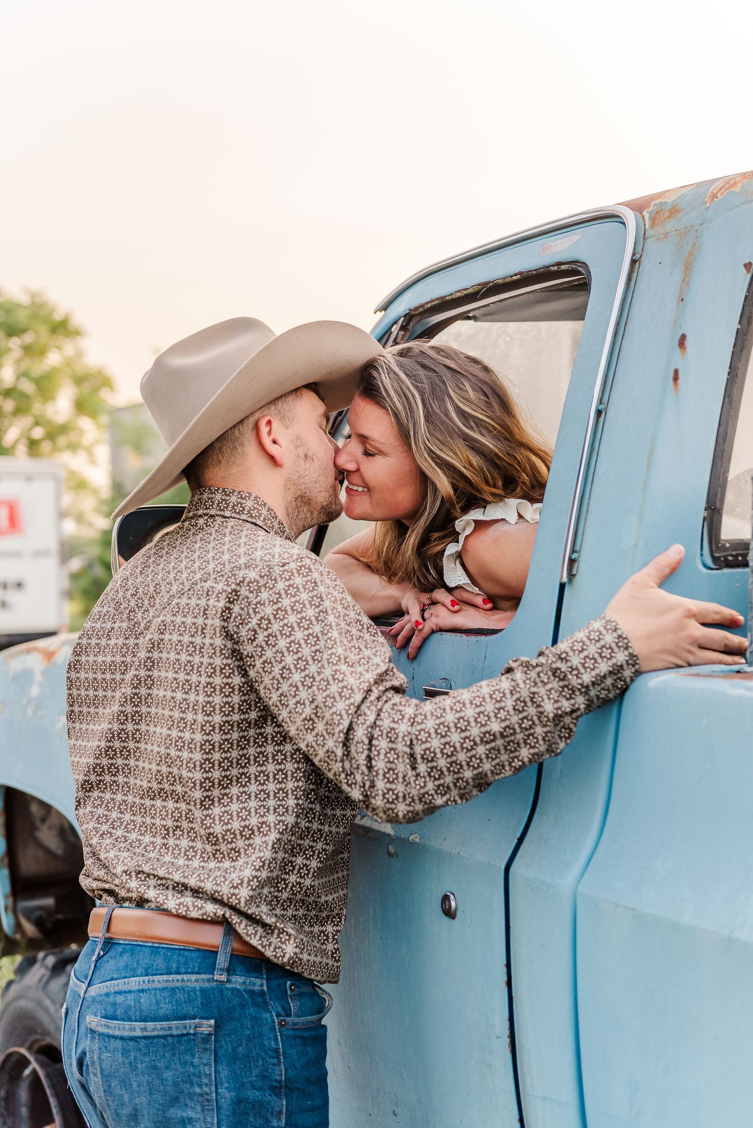 A couple shares a romantic kiss through a vintage blue truck window while wearing western attire.