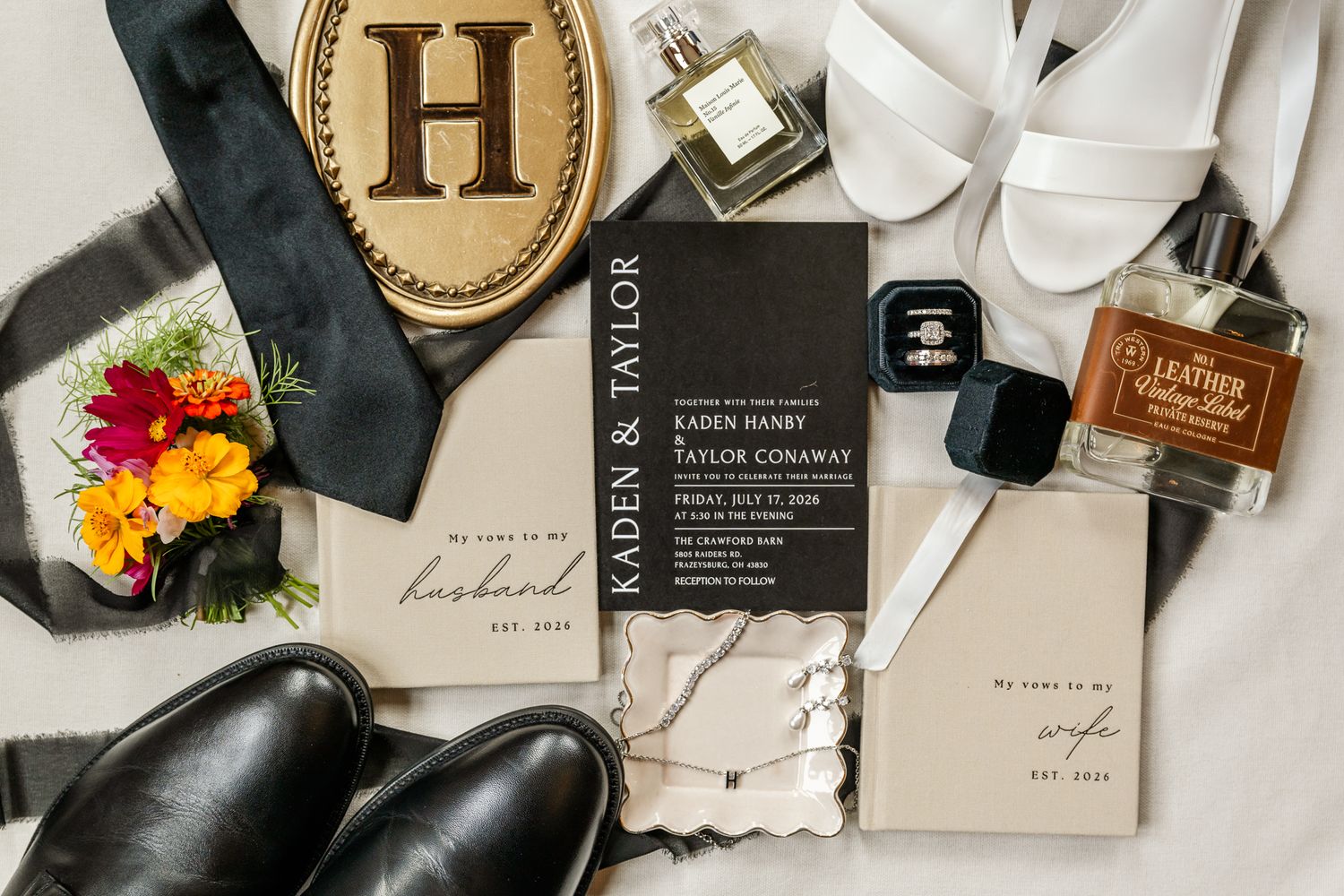 Elegant wedding flat lay featuring invitation suite, rings, cologne, black shoes, floral boutonniere, and monogram accessories.