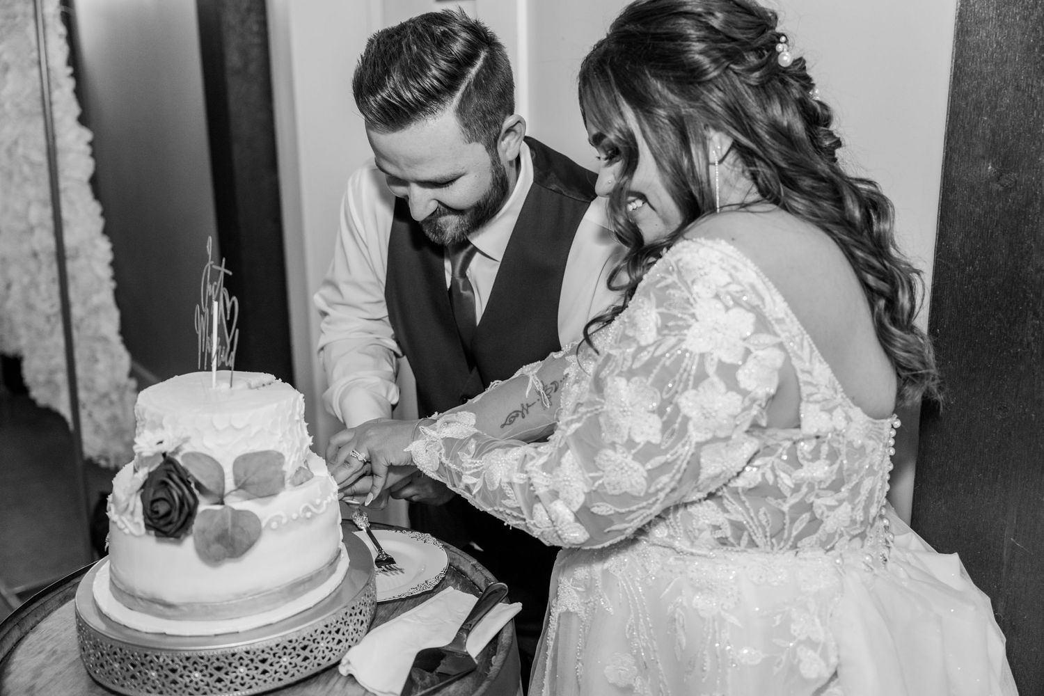 A bride and groom cut their elegant two-tier wedding cake together in a black and white photographFour Seasons Barn Wedding & Event Venue