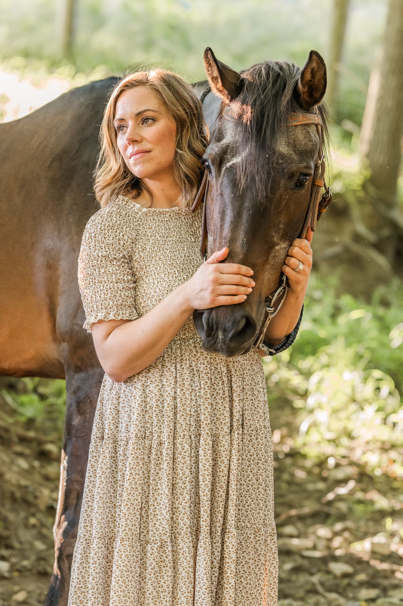 A woman in a floral dress tenderly embraces a dark brown horse in a sunlit forest setting.