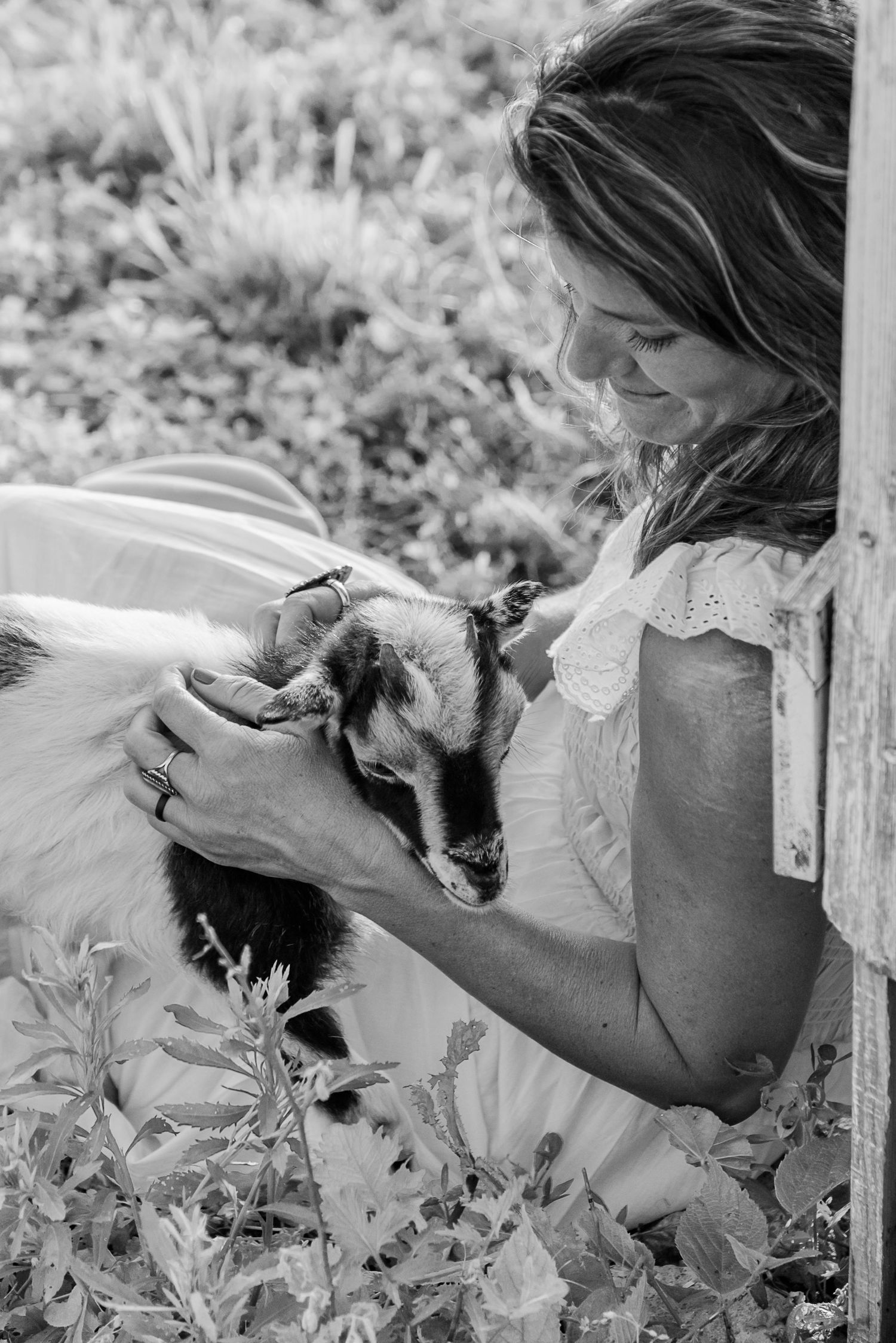 A tender moment captured in black and white as a person in a lace top gently pets a baby goat among garden foliage.