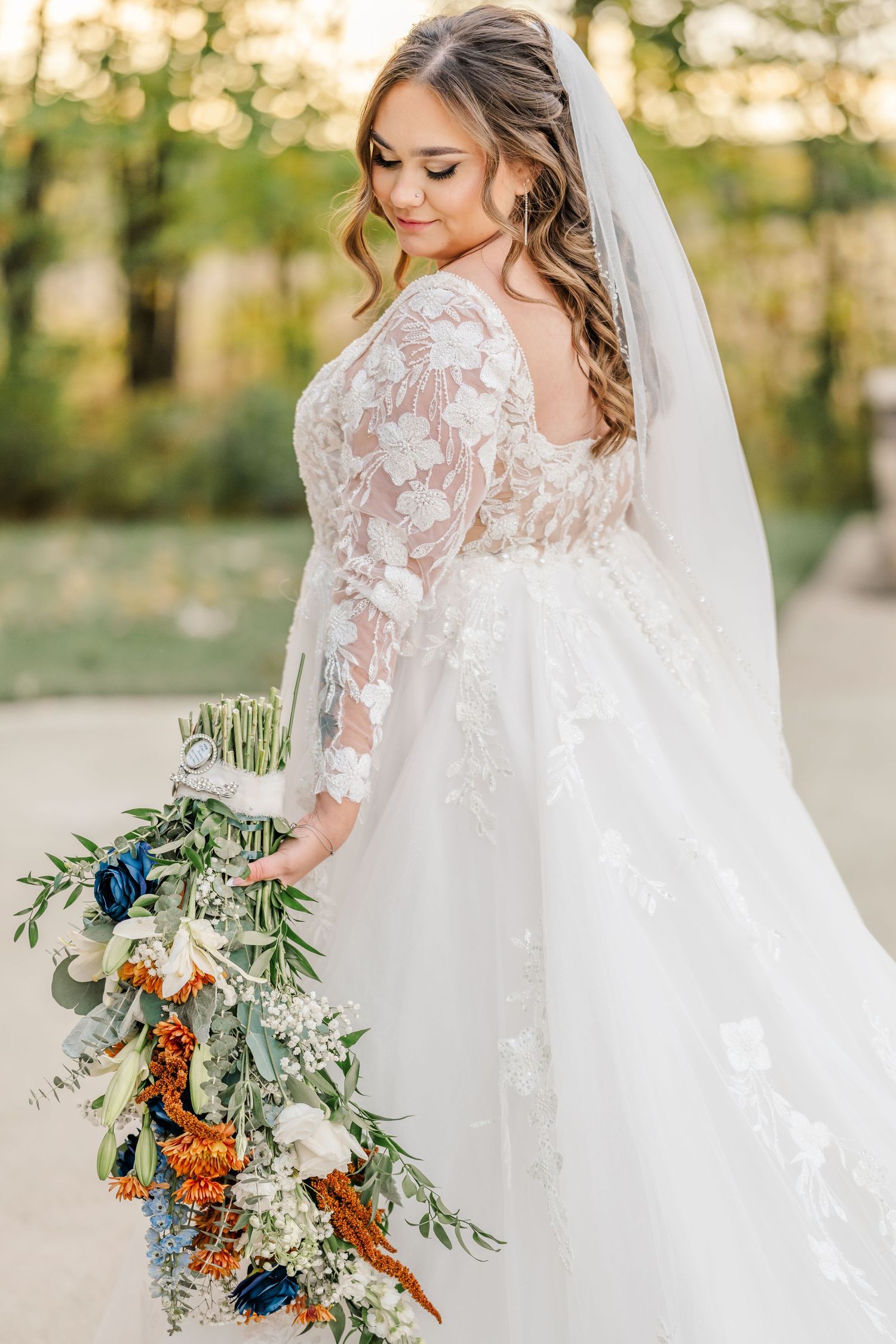 Bride in lace wedding dress holding vibrant orange and blue floral bouquet with greenery. Four Seasons Barn Wedding & Event Venue