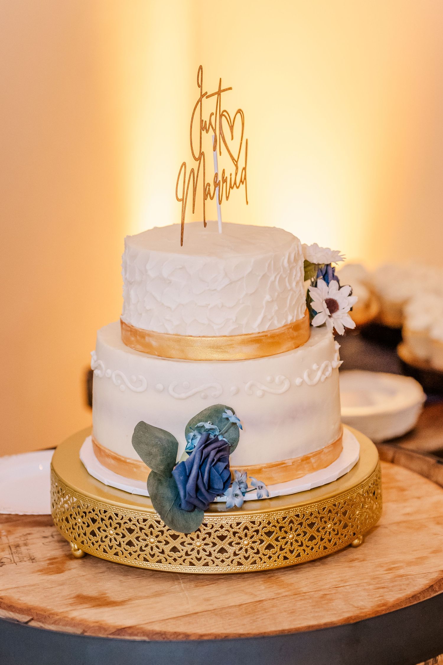 An elegant two-tier white wedding cake with gold accents and a blue flower on a decorative gold stand.