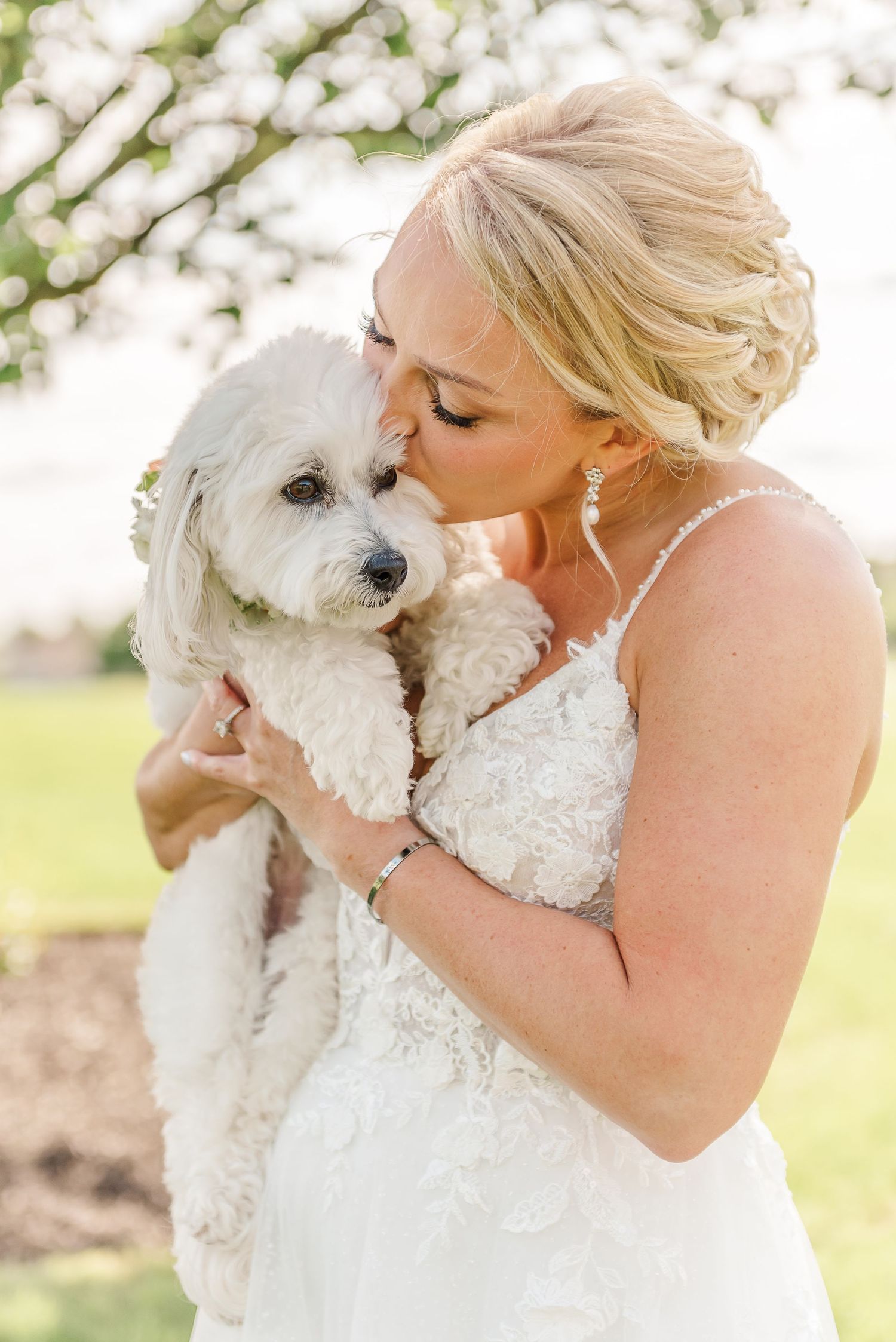 A bride in a white dress gives her small white dog a kiss during outdoor wedding portraits.