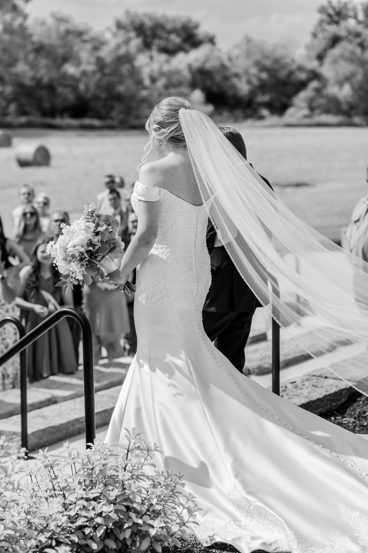 A flowing white wedding veil catches sunlight in an outdoor garden setting as it trails behind the bride.The Old Barns at Dry Run Farms