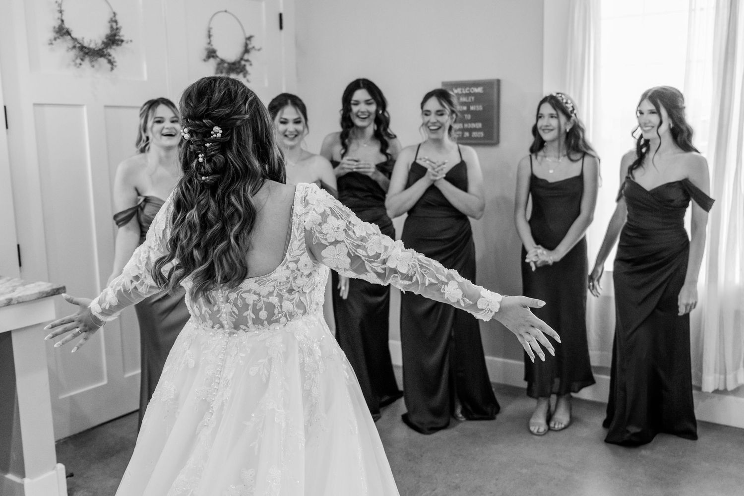 A wedding party in black dresses poses and celebrates together in a white room. Four Seasons Barn Wedding & Event Venue