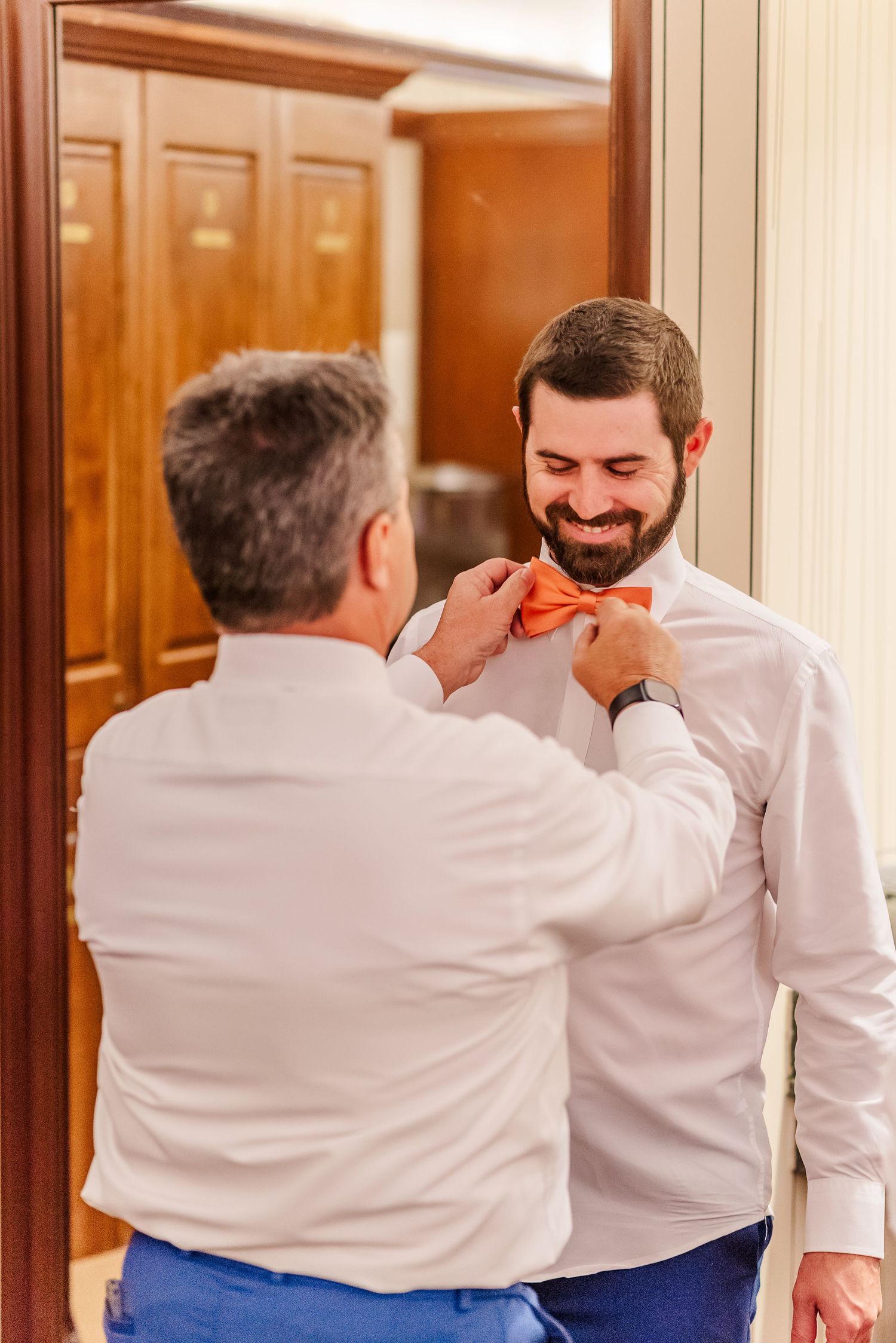 A tender moment as someone helps adjust a coral-colored bowtie on a smiling person in a white dress shirt.