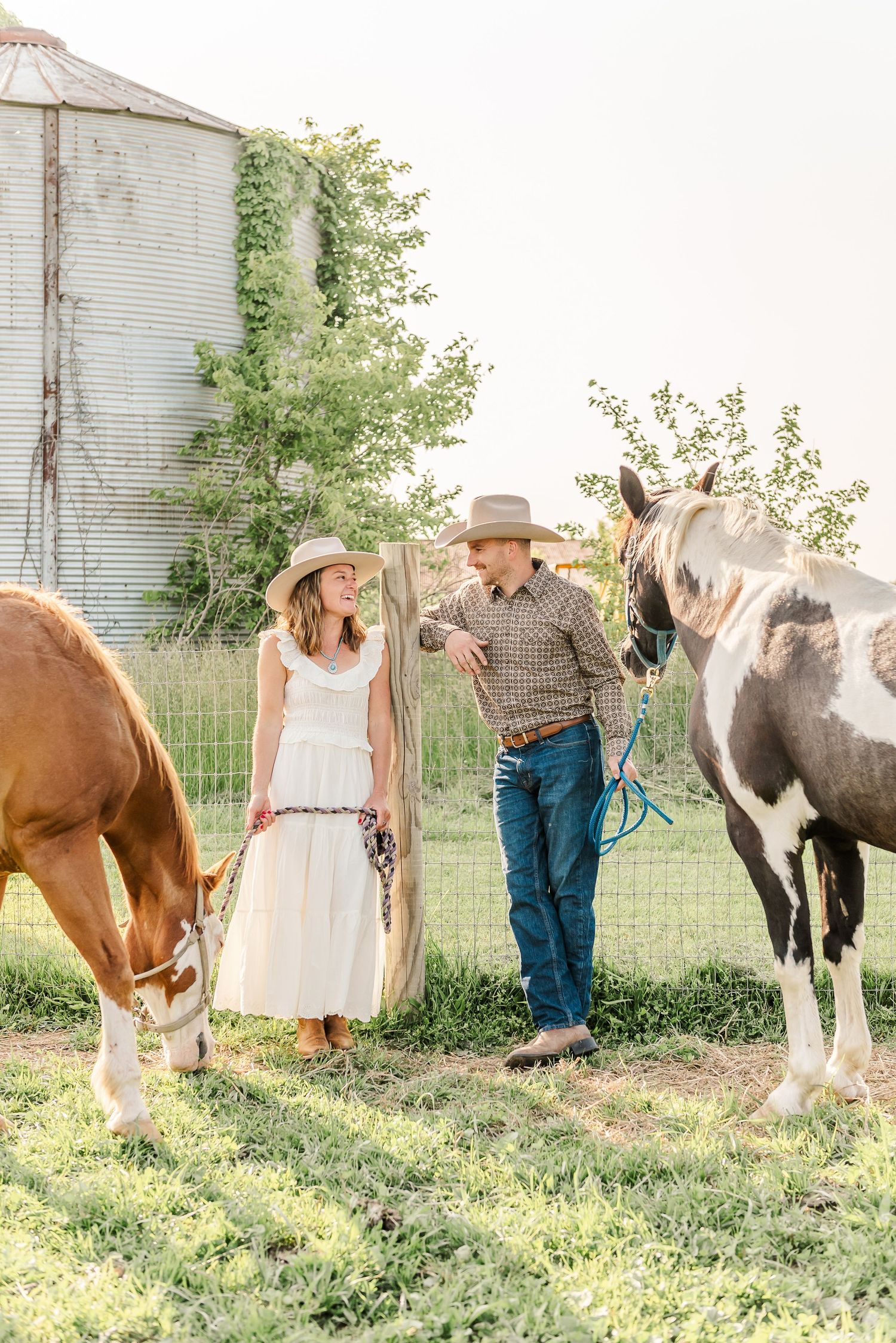 A couple in western attire stands with horses in front of old farm silos at sunset.